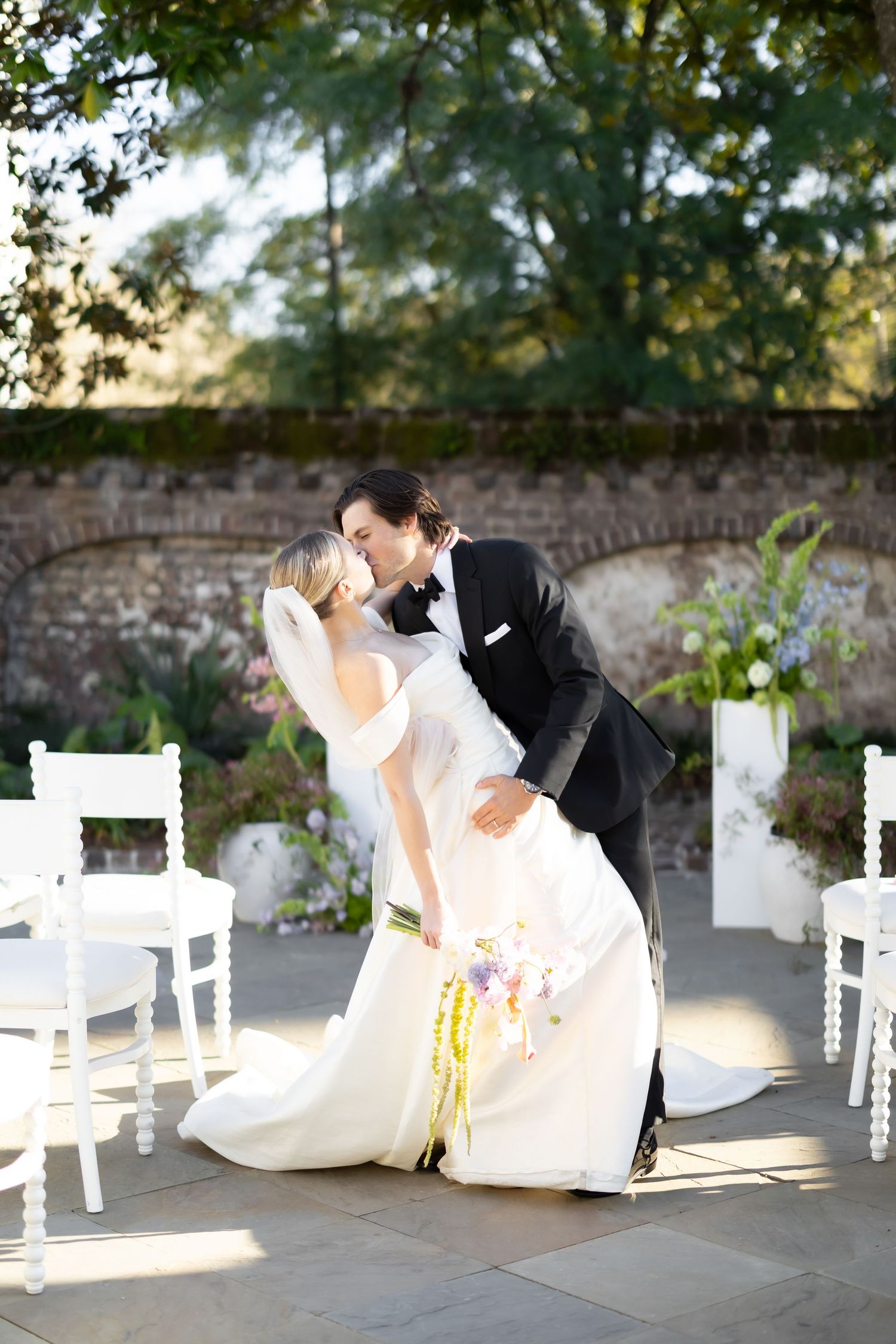 Wedding couple posing near white chairs and brick wall in garden setting.