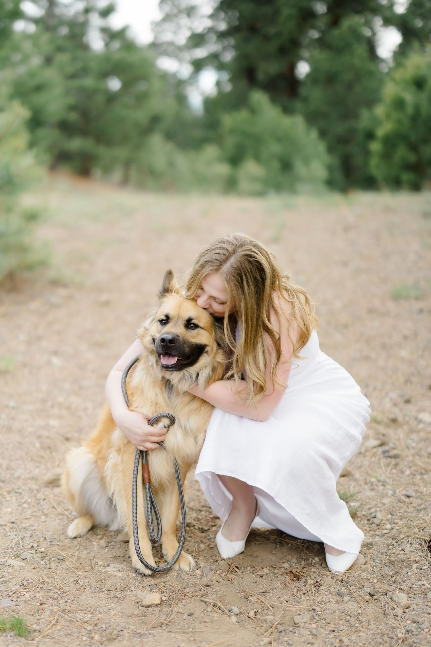 Young girl in white dress hugging a German Shepherd dog on a dirt path surrounded by green trees.