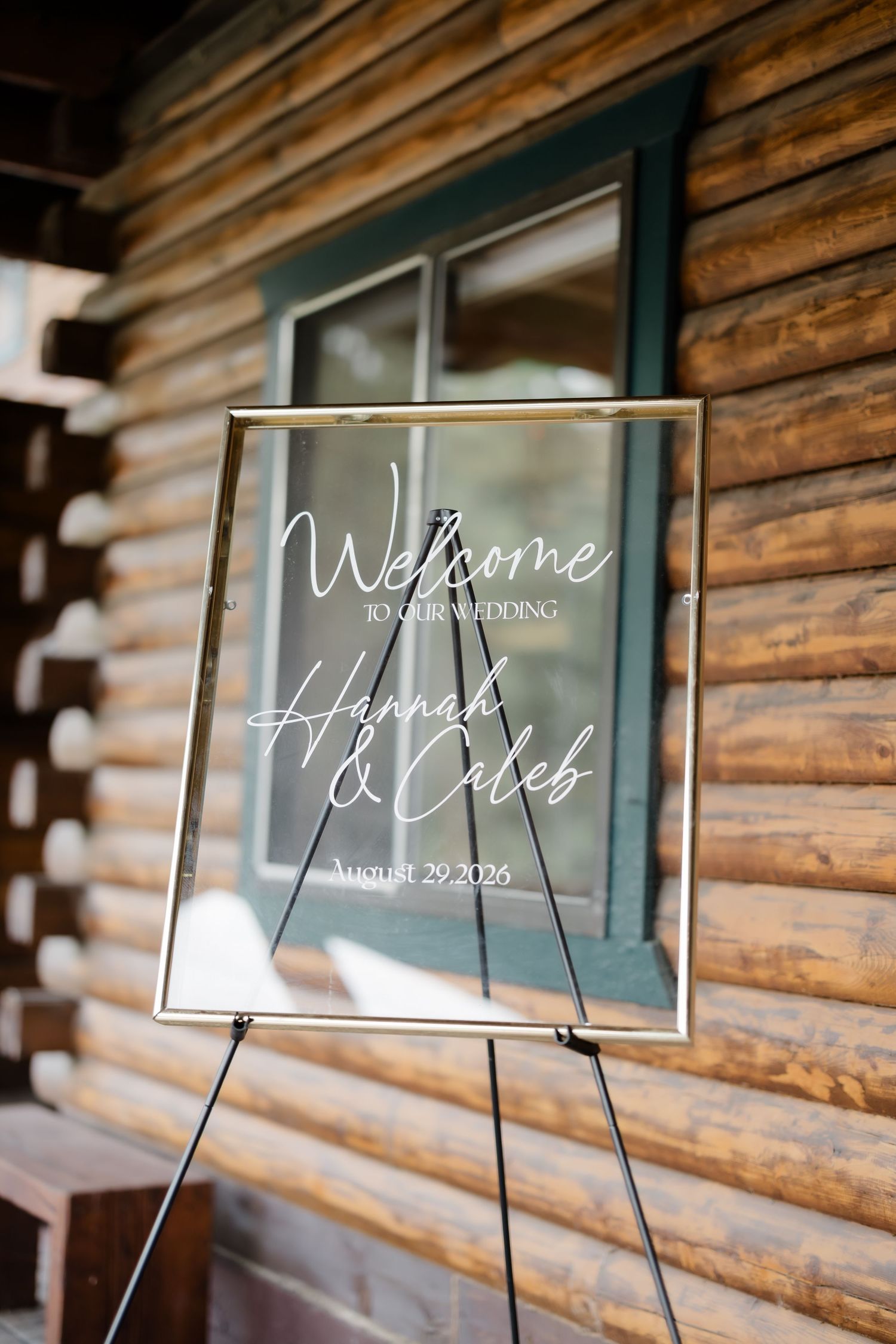 Elegant acrylic wedding welcome sign for Hannah & Caleb, August 29, 2026, displayed on easel outside rustic log cabin.
