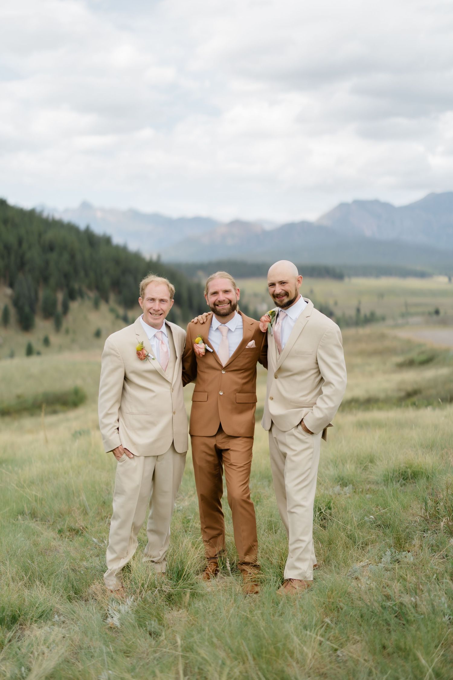 Groom in brown suit flanked by two groomsmen in tan suits, posing in a mountain meadow at an outdoor wedding.
