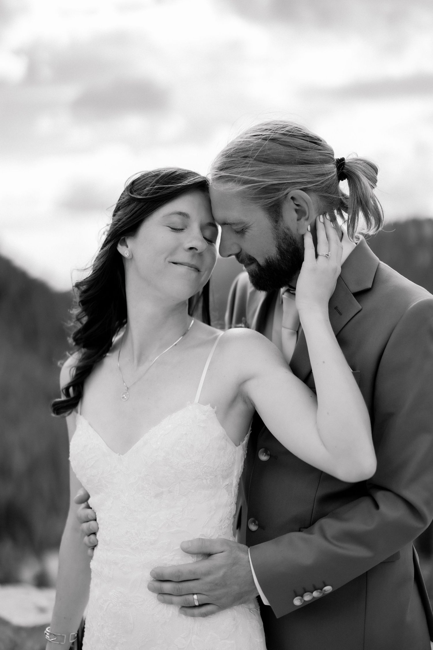 Black and white wedding photo of bride and groom touching foreheads tenderly outdoors with mountains in background.