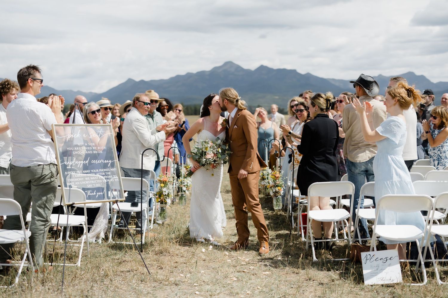 Bride and groom share a kiss at outdoor mountain wedding ceremony with guests applauding along the aisle.