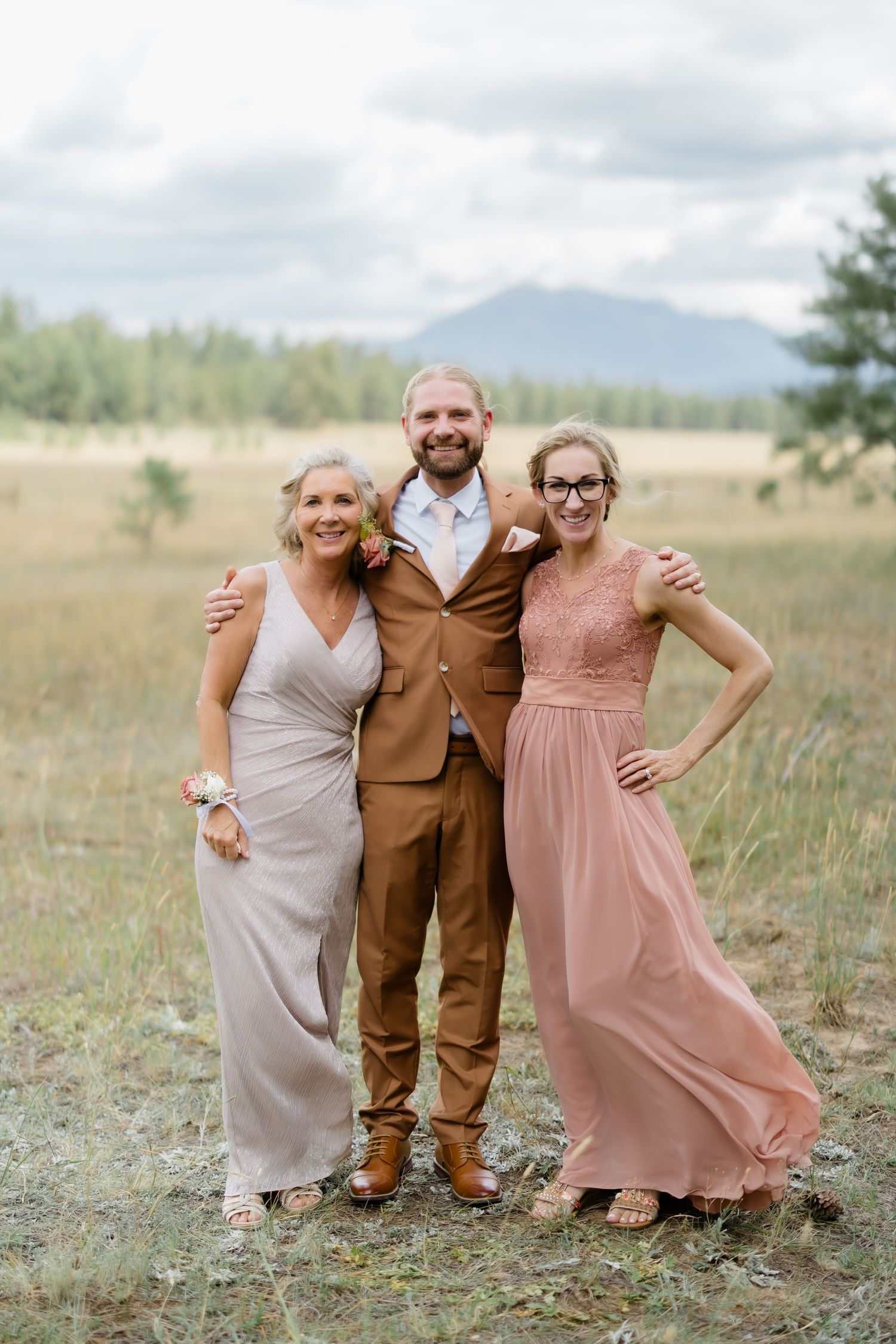A man in a brown suit poses with two women in formal dresses at an outdoor wedding in a mountain meadow.