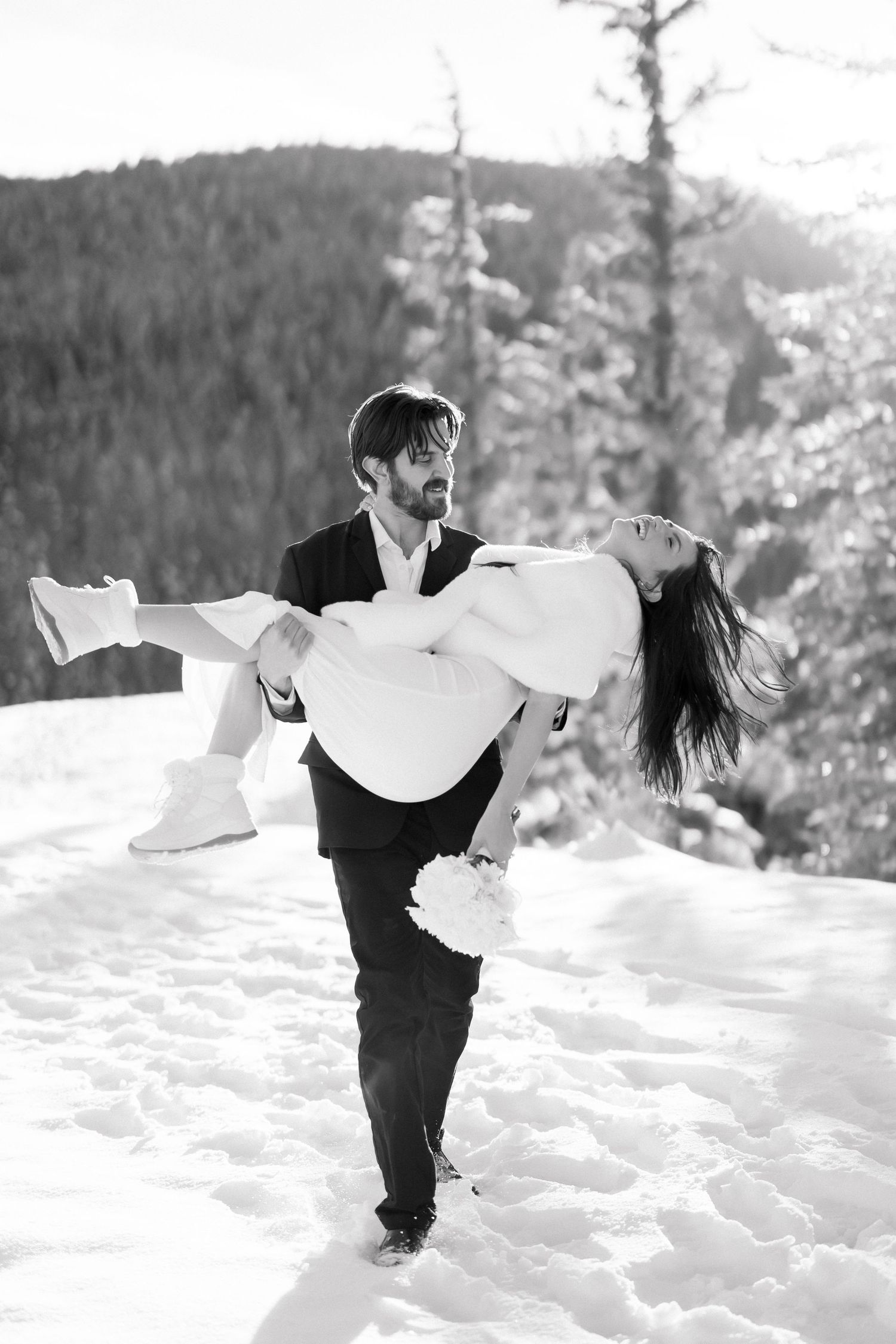 Black and white photo of groom carrying bride in his arms through snowy mountain landscape.