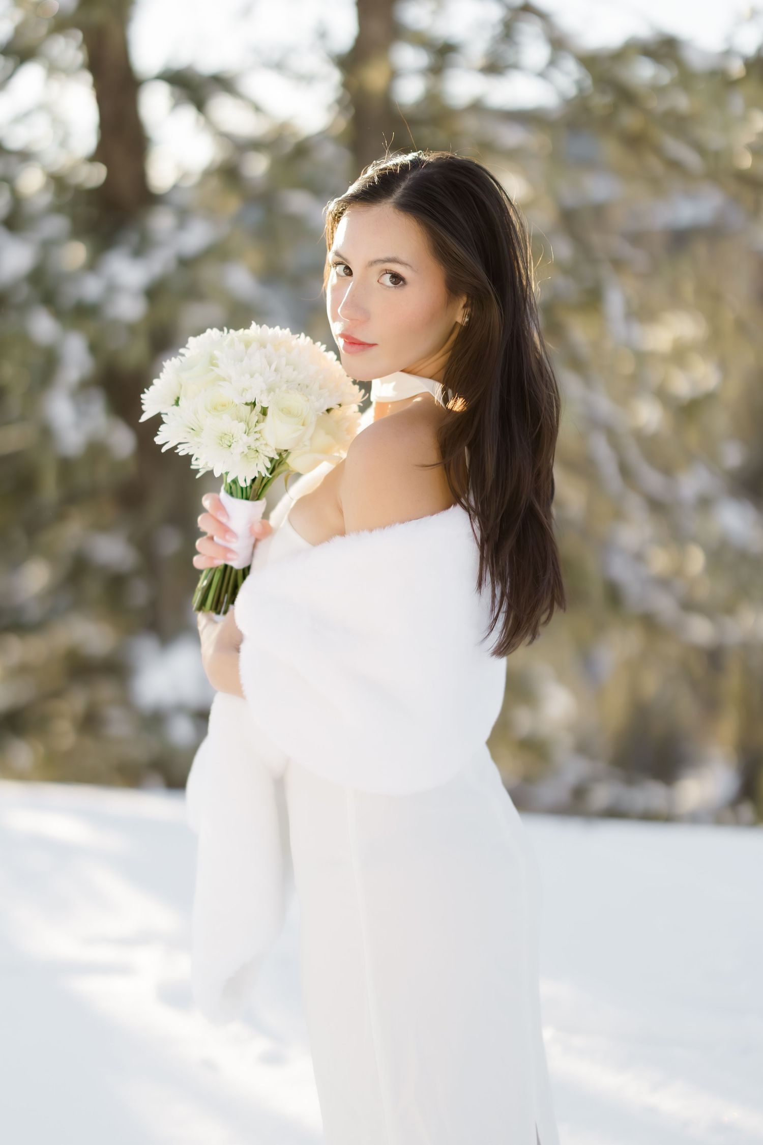 Elegant bride in white dress holding bouquet of white flowers in snowy winter outdoor setting.