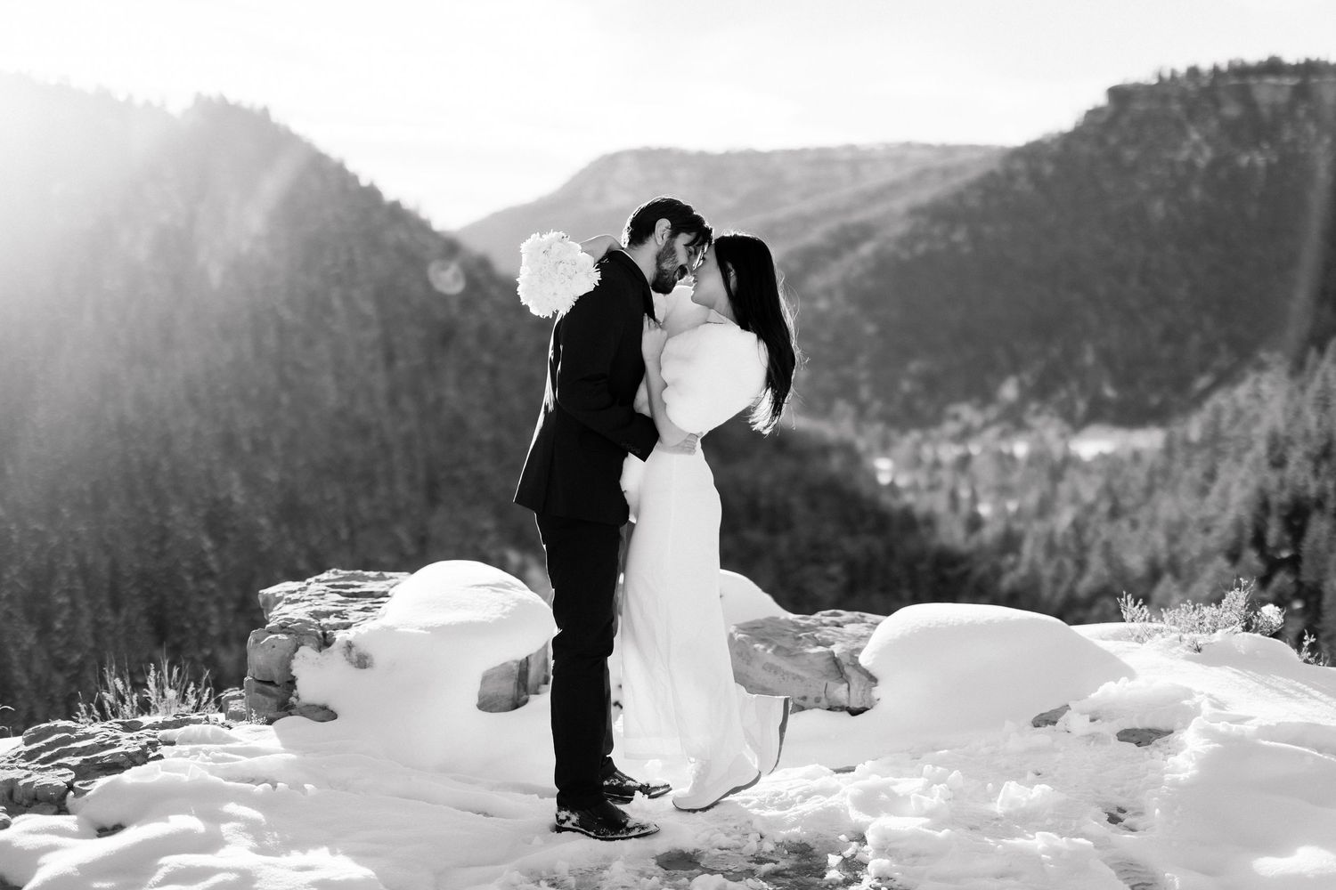 Black and white photo of bride and groom embracing on snowy mountain peak with forest backdrop.