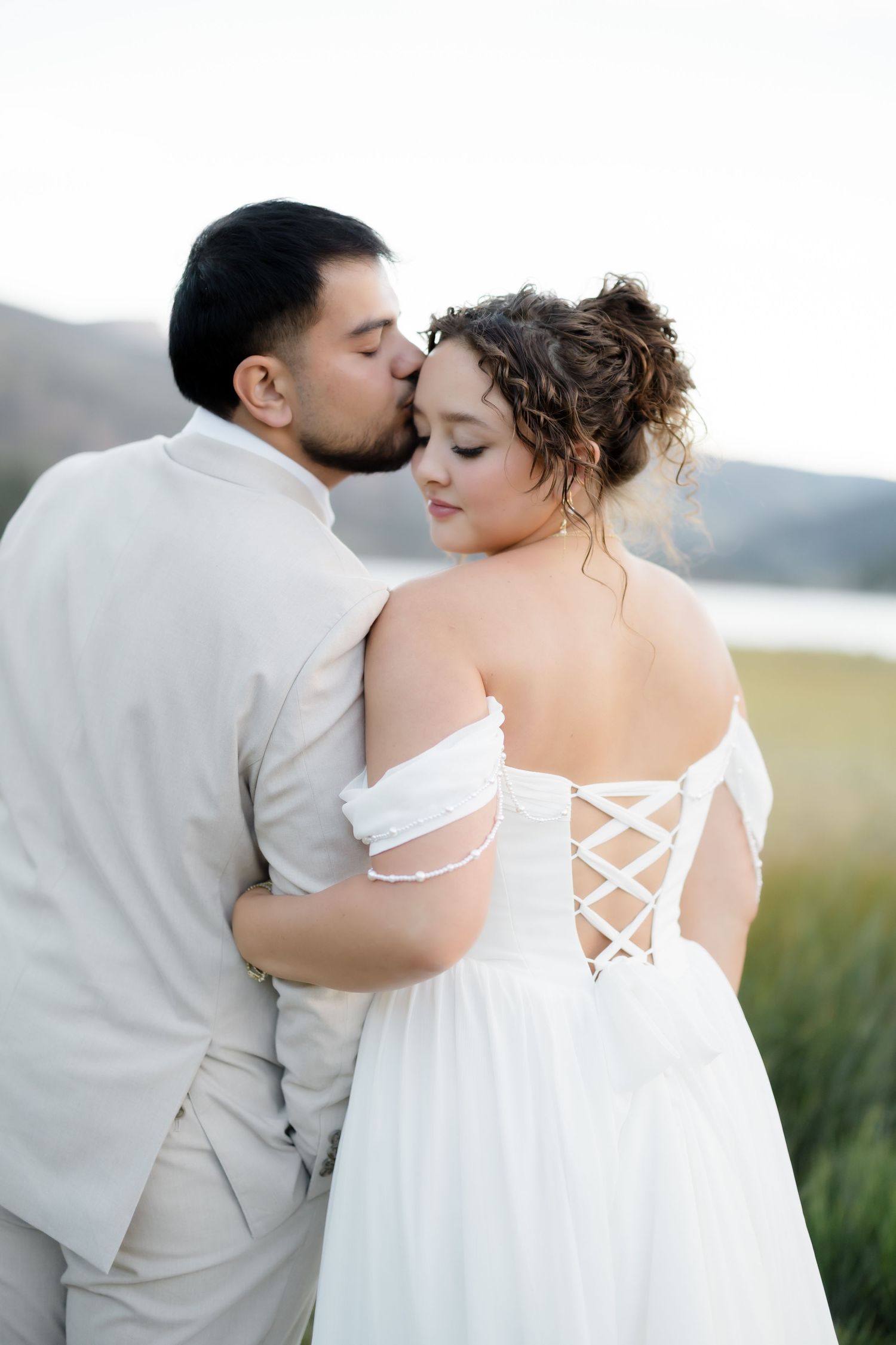 Groom kisses bride on cheek from behind, showcasing the corset lace-up back of her white off-shoulder gown.