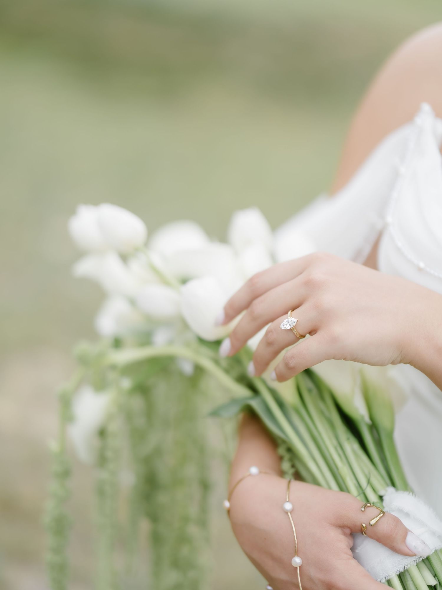 Bride holding white tulip bouquet with gold engagement ring and pearl bracelet in soft outdoor setting.