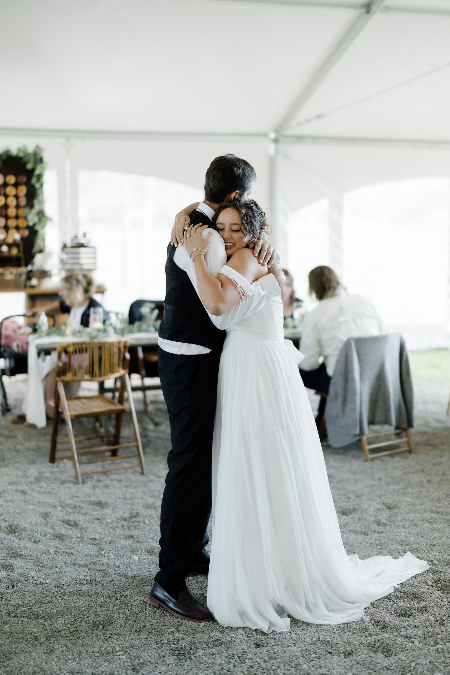 A bride in a flowing white gown shares a tender first dance embrace with her groom under a white wedding reception tent.