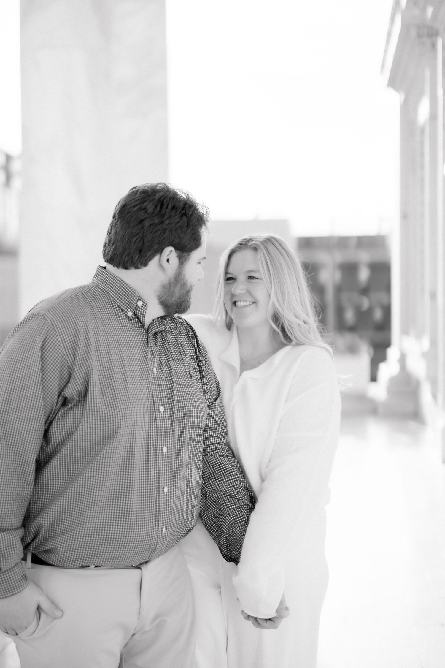Black and white portrait of a smiling woman and bearded man sharing a romantic moment by marble columns.