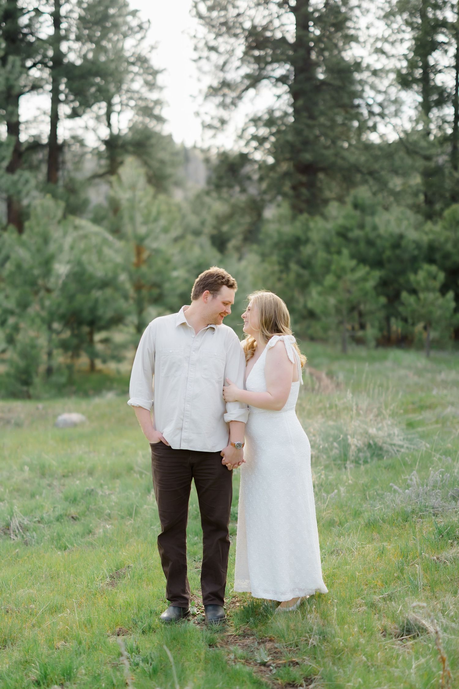 Couple holding hands and smiling at each other in a lush green meadow surrounded by pine trees.