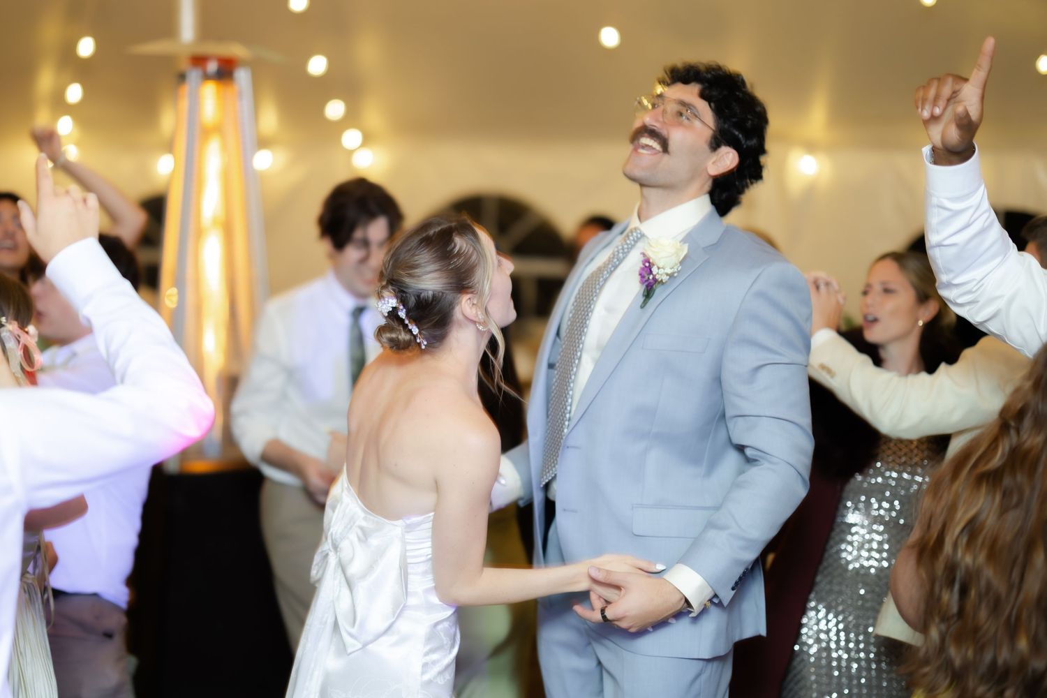 Bride and groom dancing joyfully at wedding reception under warm string lights with guests celebrating.