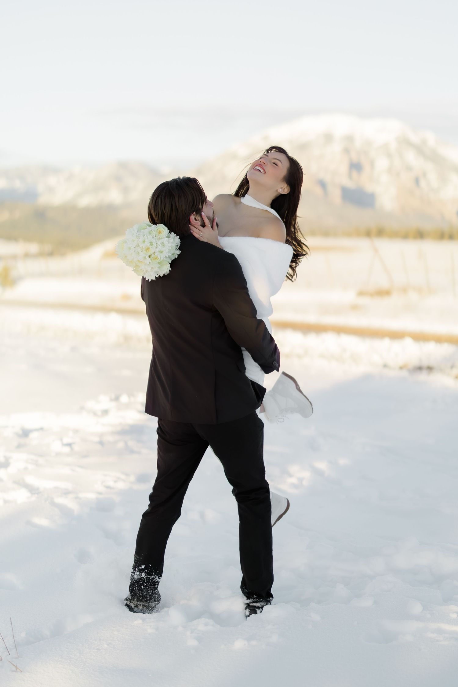 Groom lifts bride in white dress and fur stole, spinning her joyfully in a snowy mountain landscape.