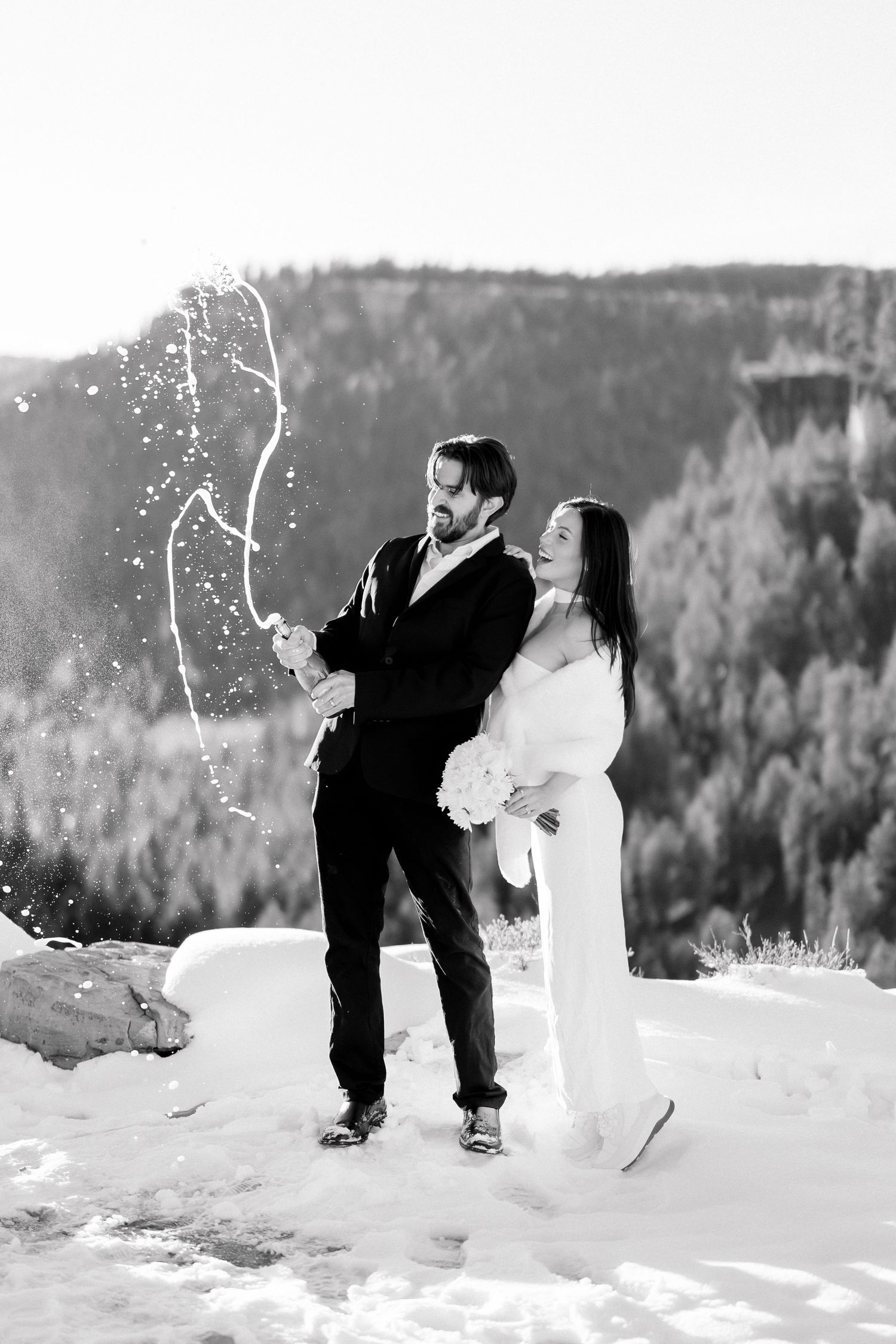 Black and white photo of couple popping champagne in snow with mountain forest backdrop.