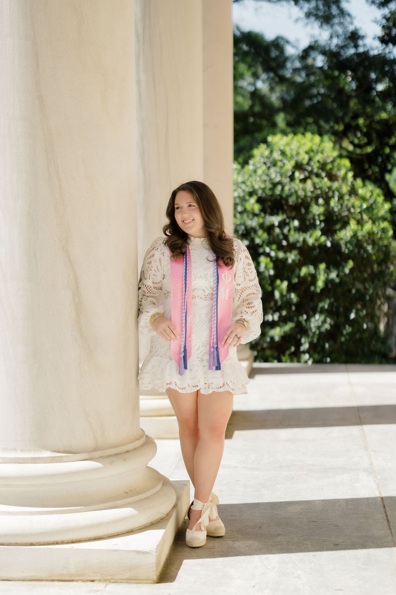 Smiling young woman in white lace dress with colorful graduation cords leaning against marble column outdoors.
