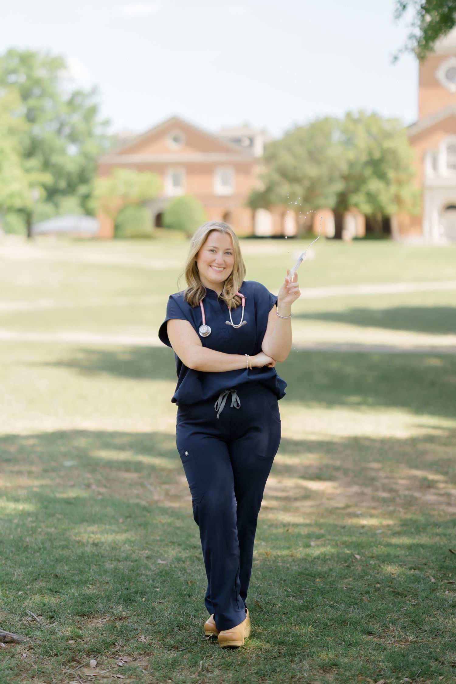 Smiling female nurse in navy scrubs with stethoscope posing outdoors on green campus lawn near brick building.