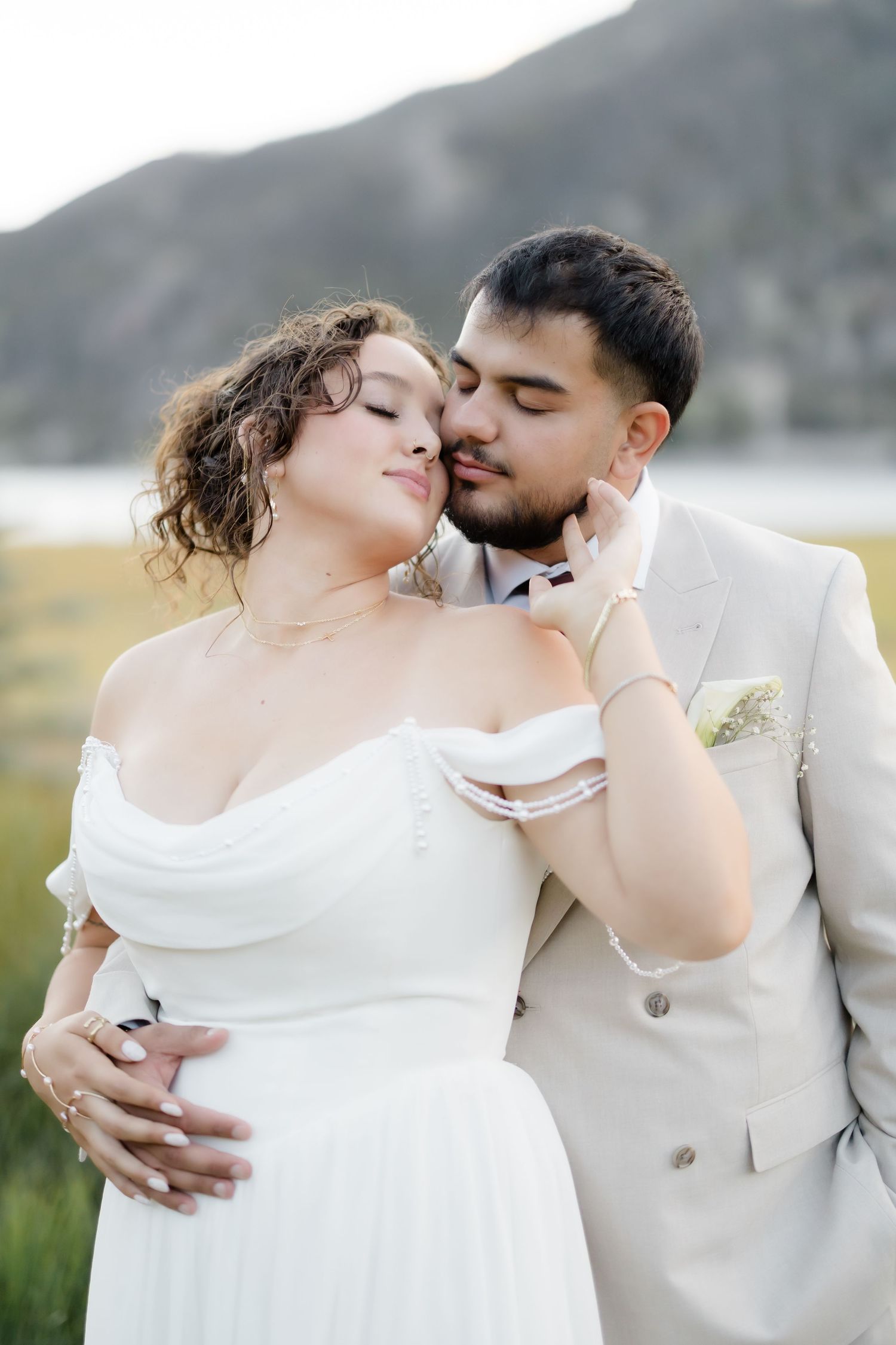 A bride and groom share a tender moment outdoors, with mountains and a lake in the background on their wedding day.