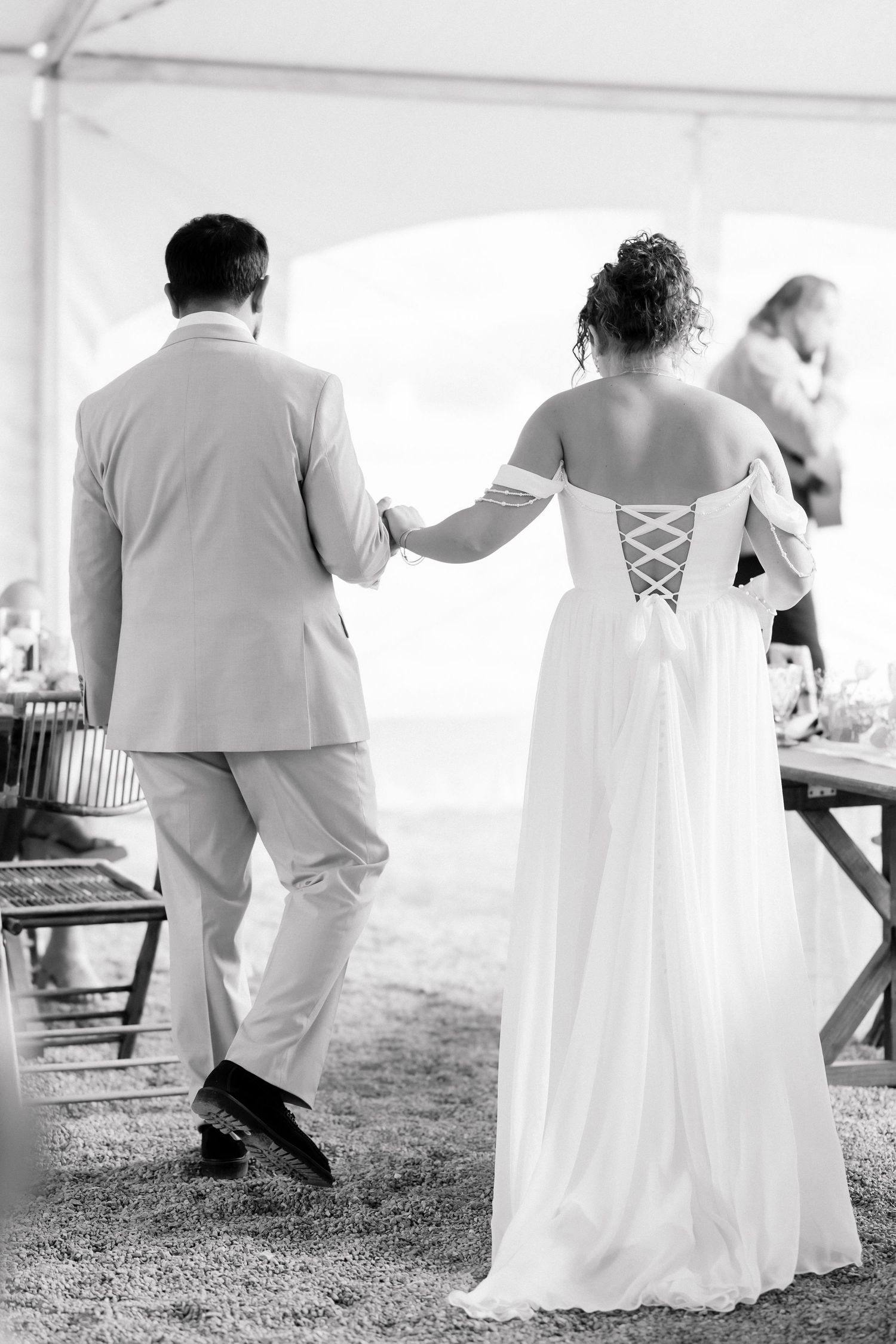 Black and white photo of a bride and groom walking hand in hand at an outdoor wedding reception.