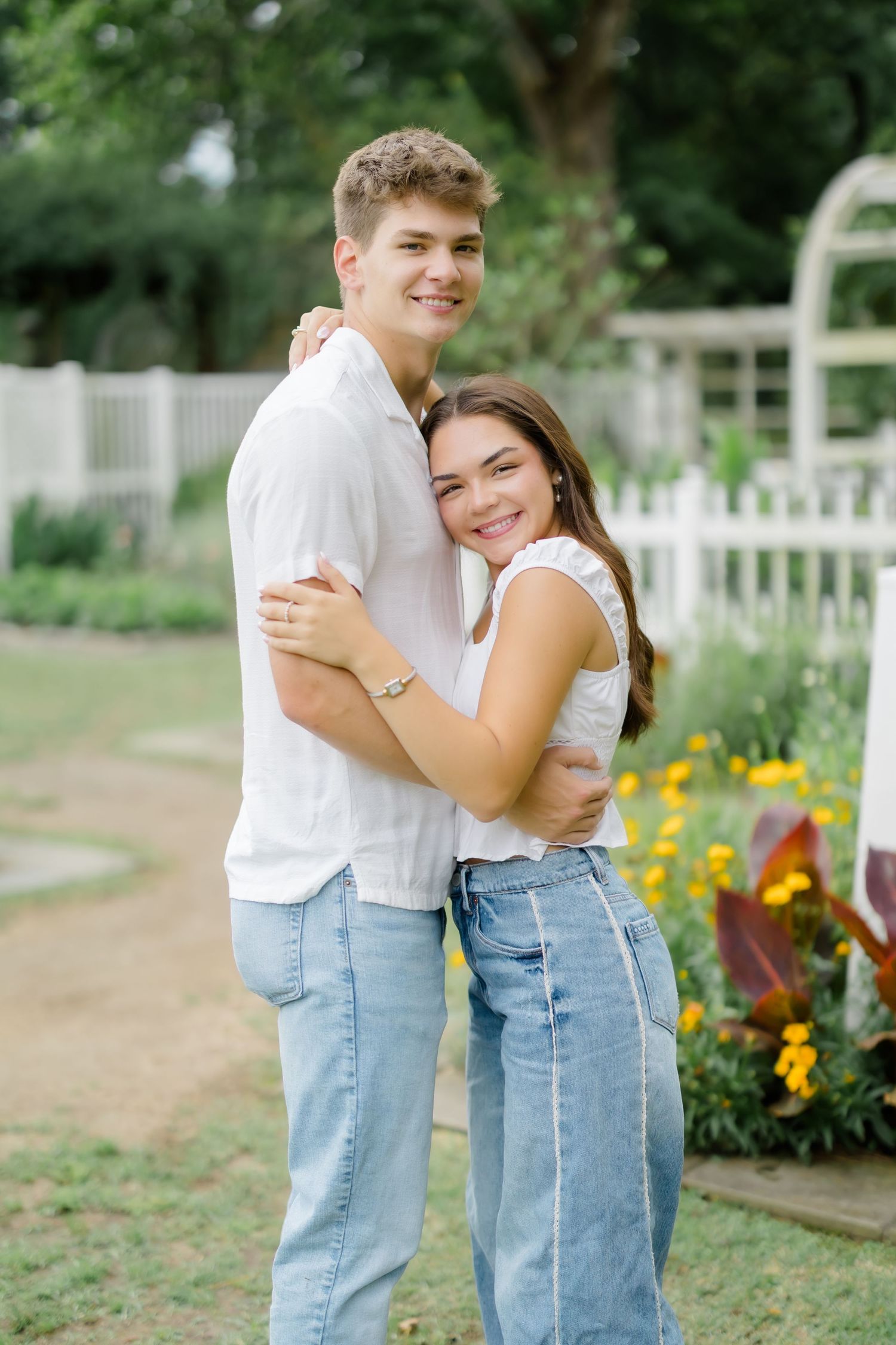 A smiling young couple embracing in a garden, wearing matching white tops and jeans near yellow flowers.