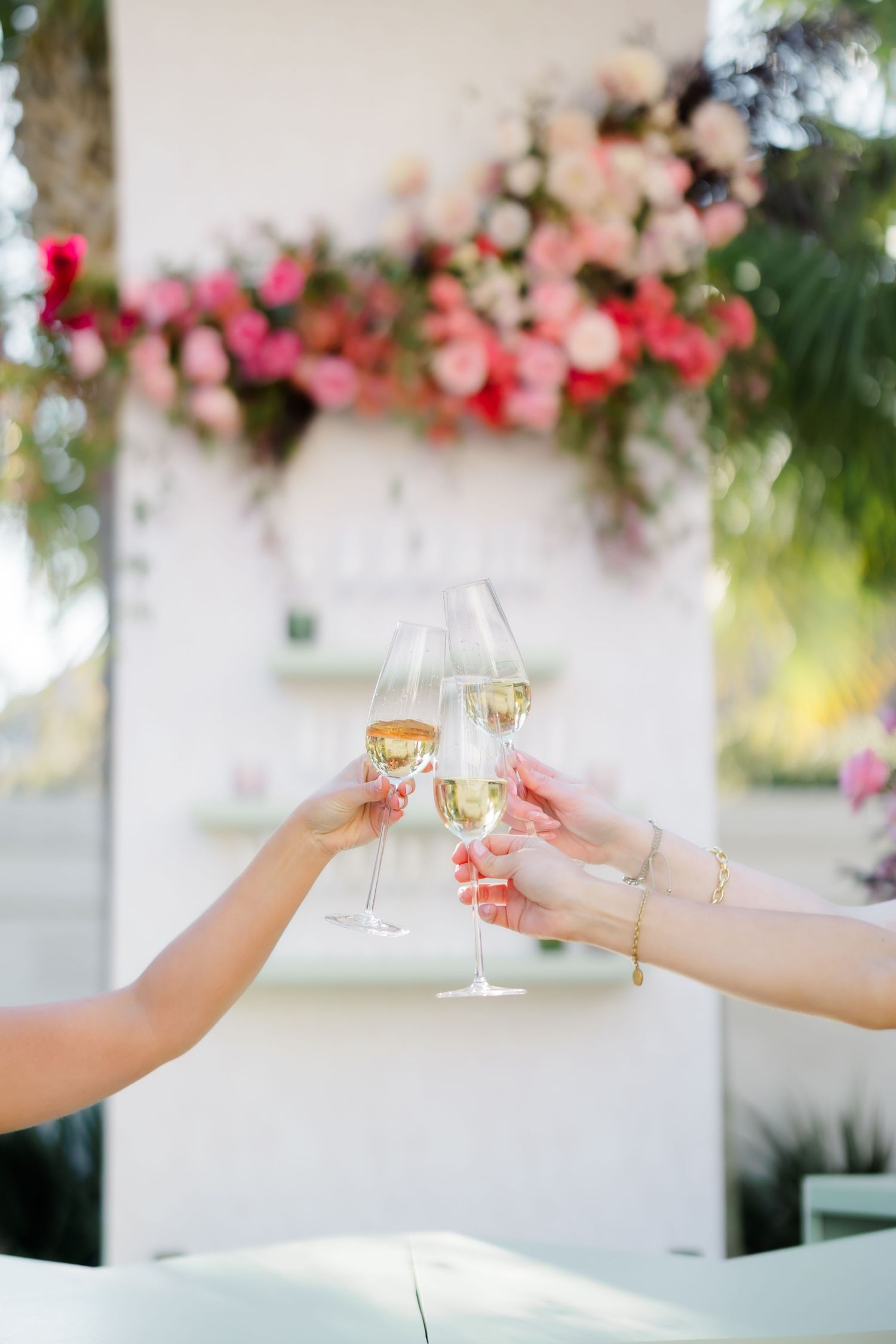 Two women toasting with champagne flutes at an outdoor celebration with pink floral arrangements in background.