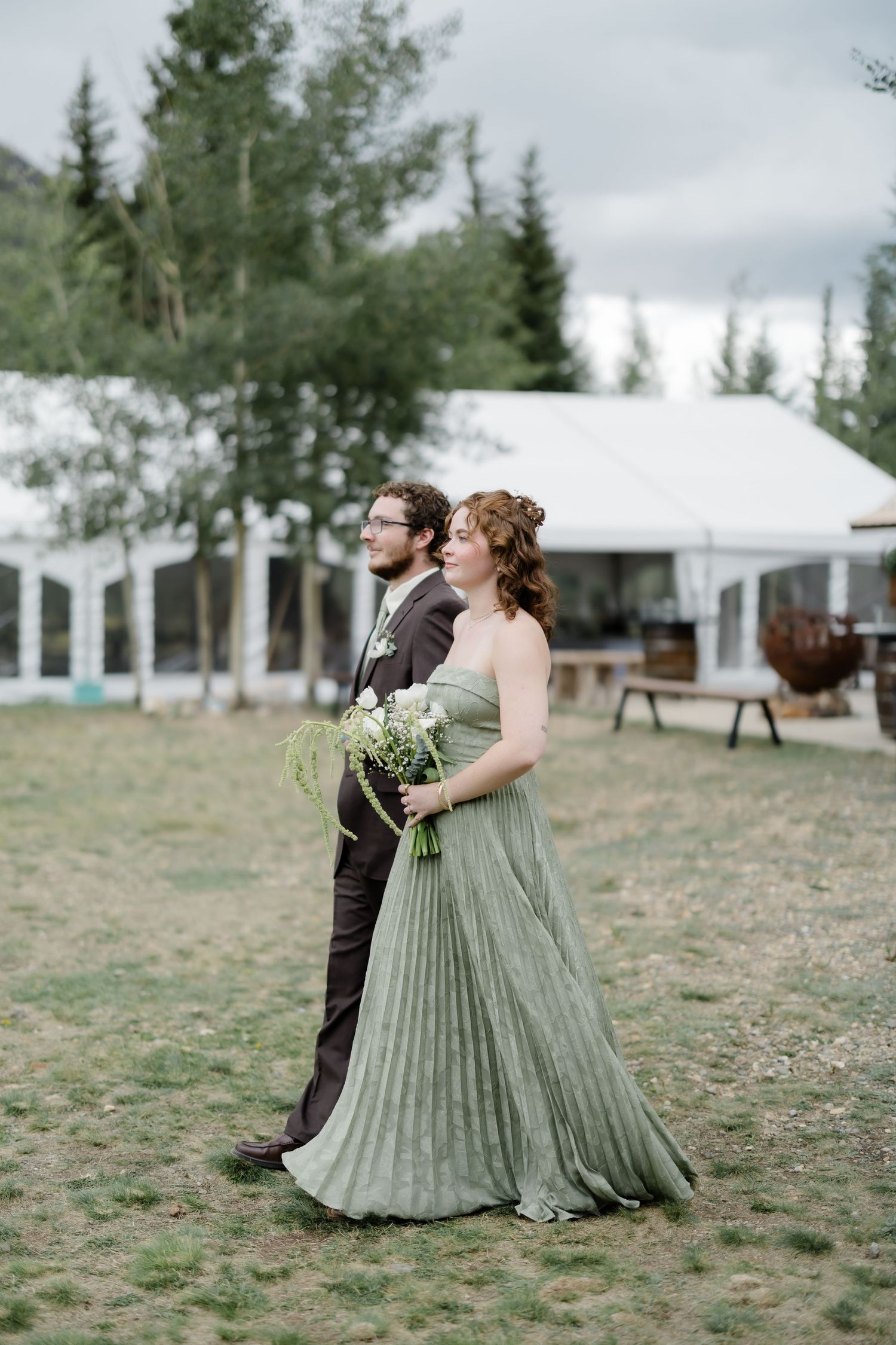 Bridesmaid in sage green pleated gown poses with groomsman at outdoor mountain wedding venue.