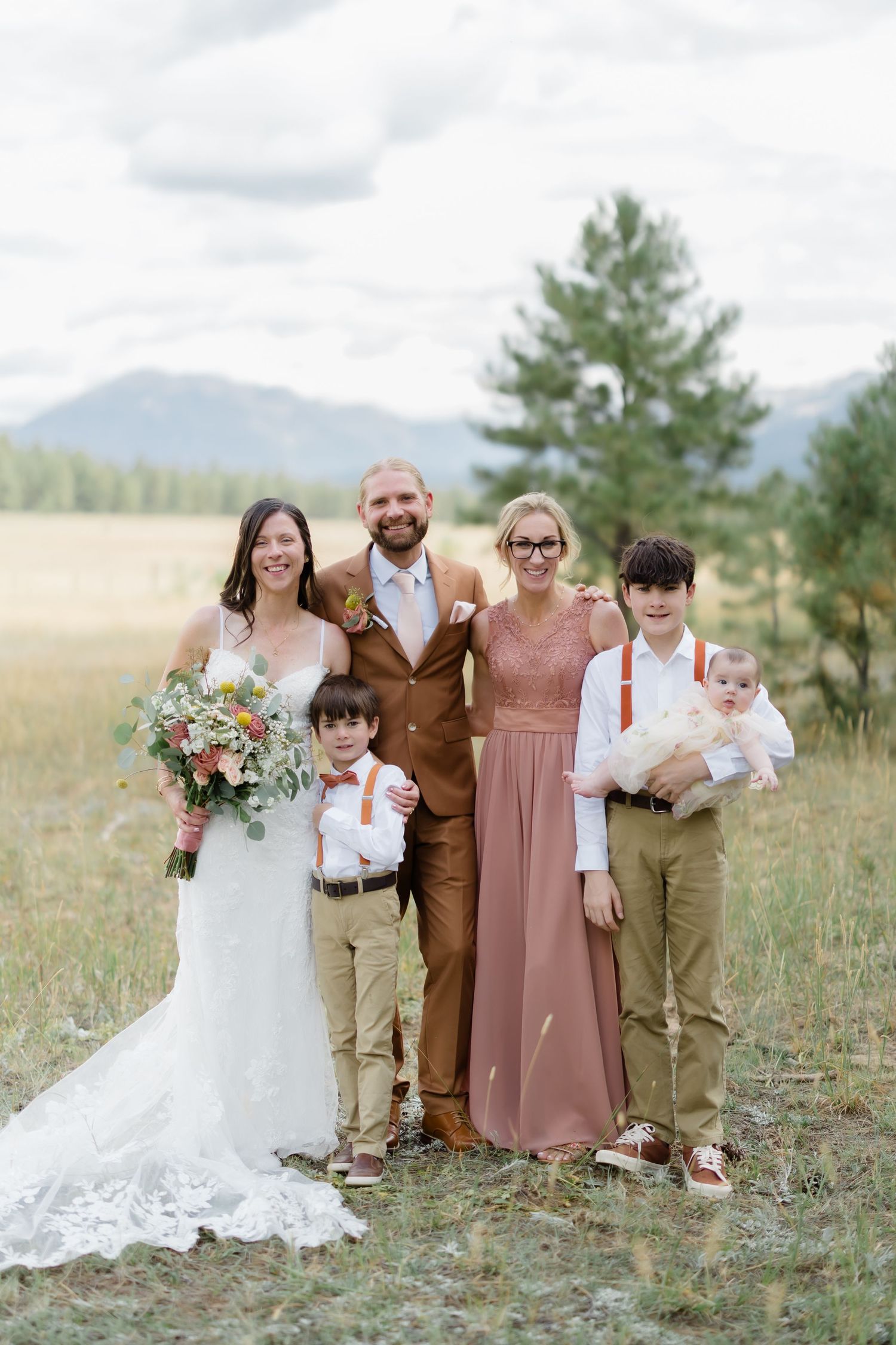 Wedding party poses outdoors in a mountain meadow, bride in white gown with bouquet beside groom and family in earthy tones.