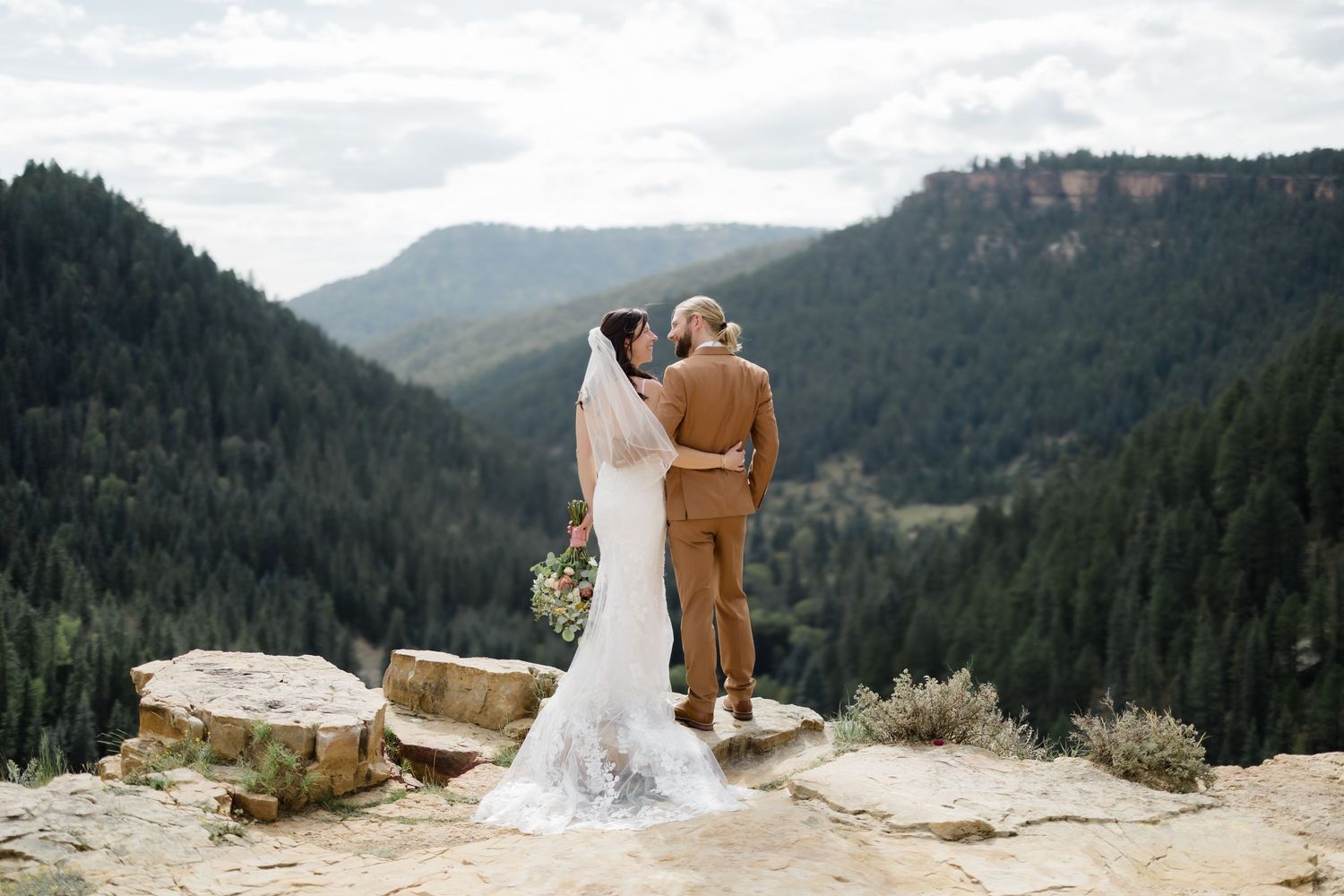 Bride and groom stand on rocky mountain overlook surrounded by pine forests, sharing an intimate moment in wedding attire.