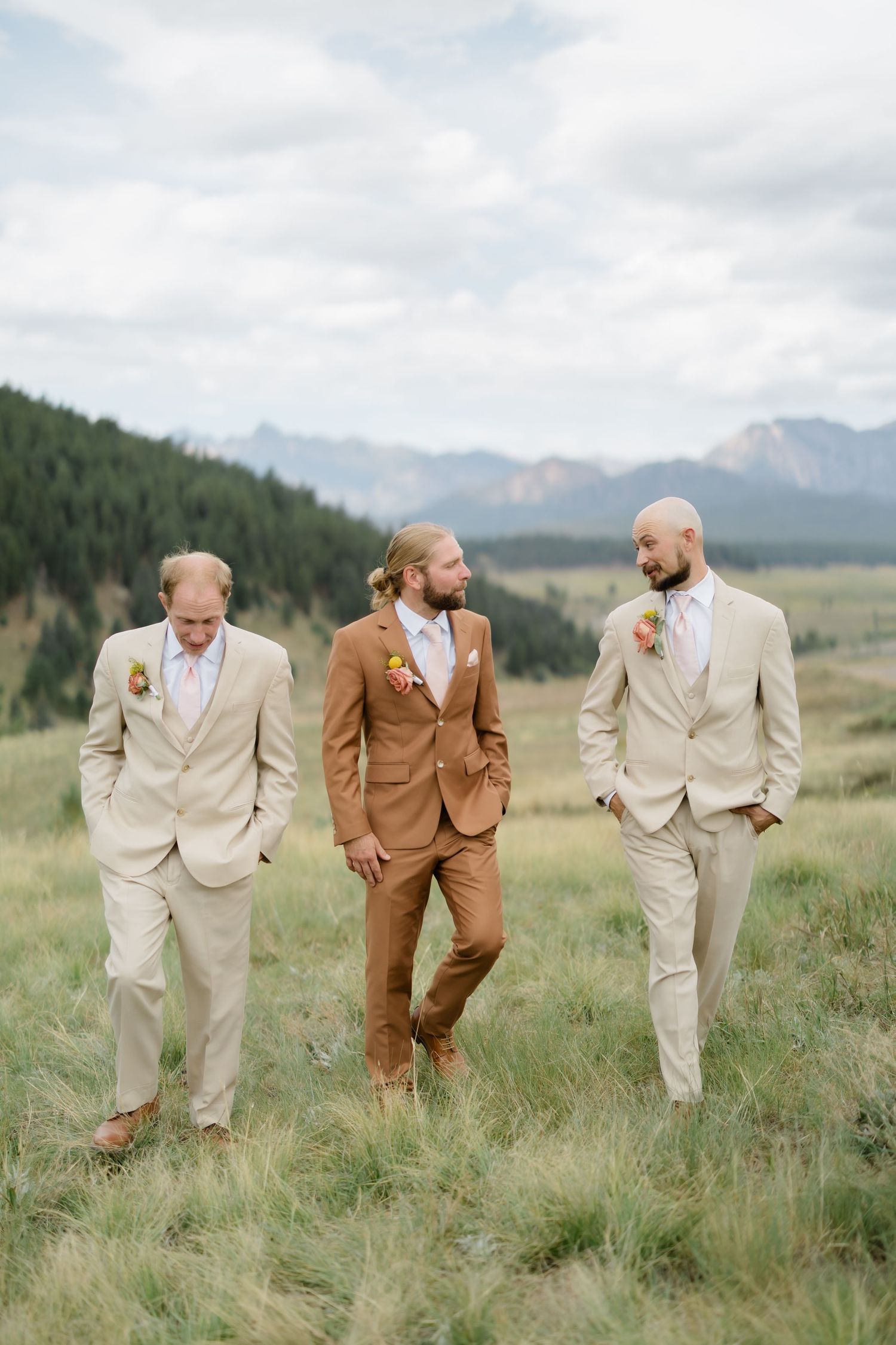 Groom in brown suit walks with two groomsmen in tan suits across a mountain meadow on a cloudy wedding day.