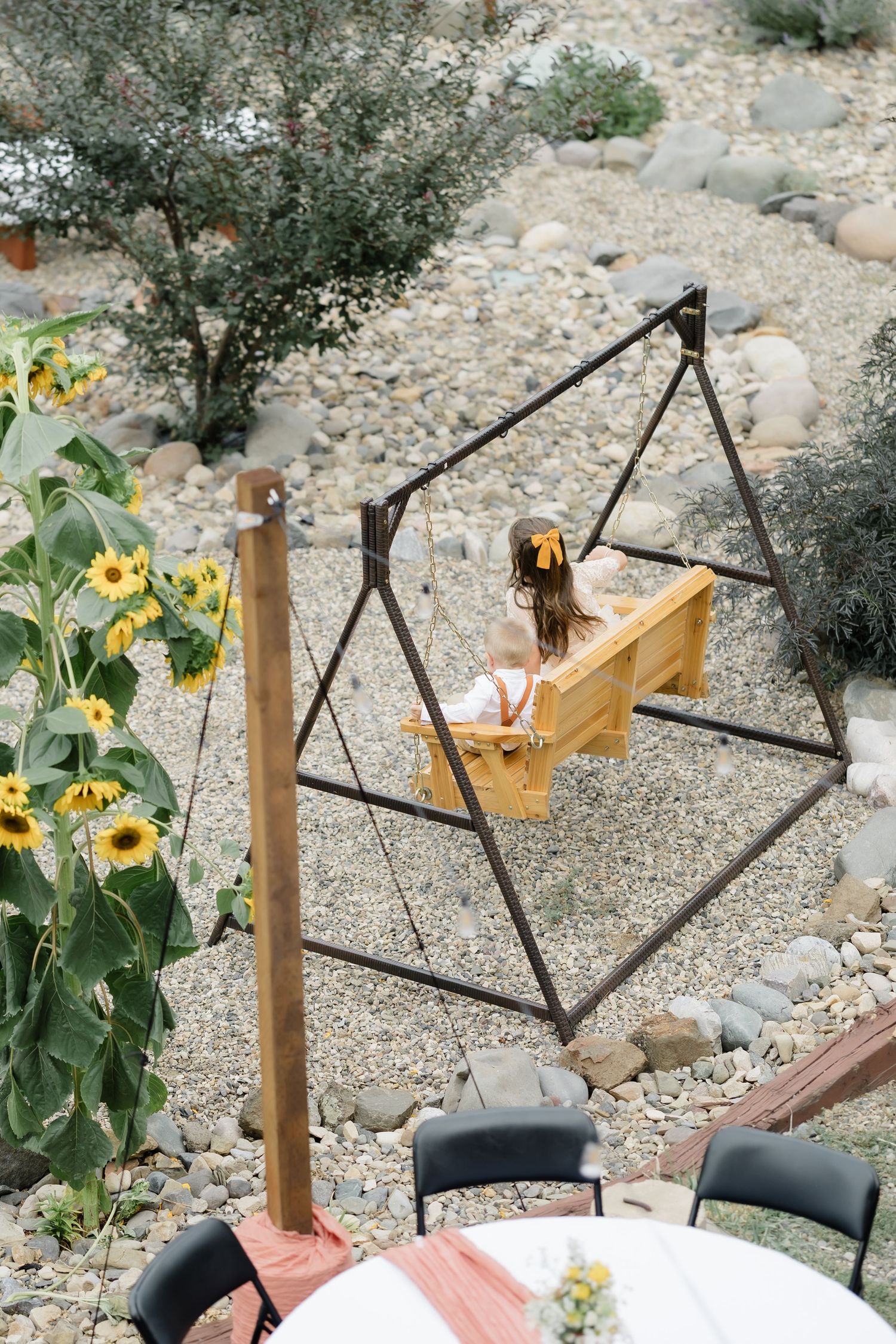 A child sits on a wooden swing set surrounded by sunflowers and a rocky garden landscape.