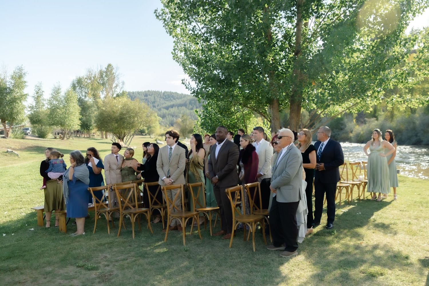 Outdoor wedding ceremony guests standing on green grass near tall trees by a river on a sunny day.