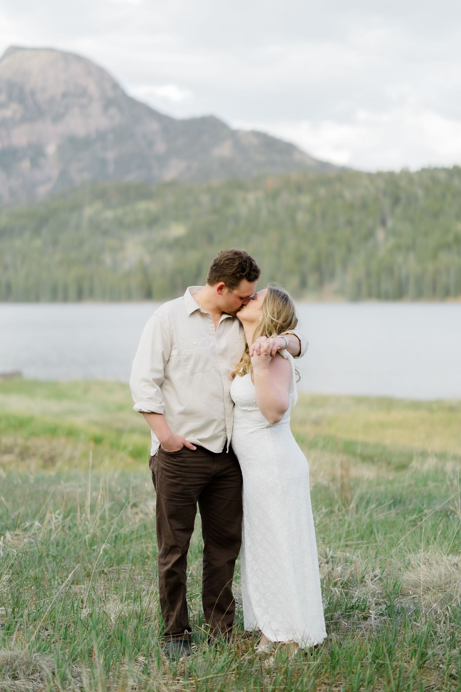 Couple sharing a romantic kiss in a meadow with a mountain lake and peaks in the background.