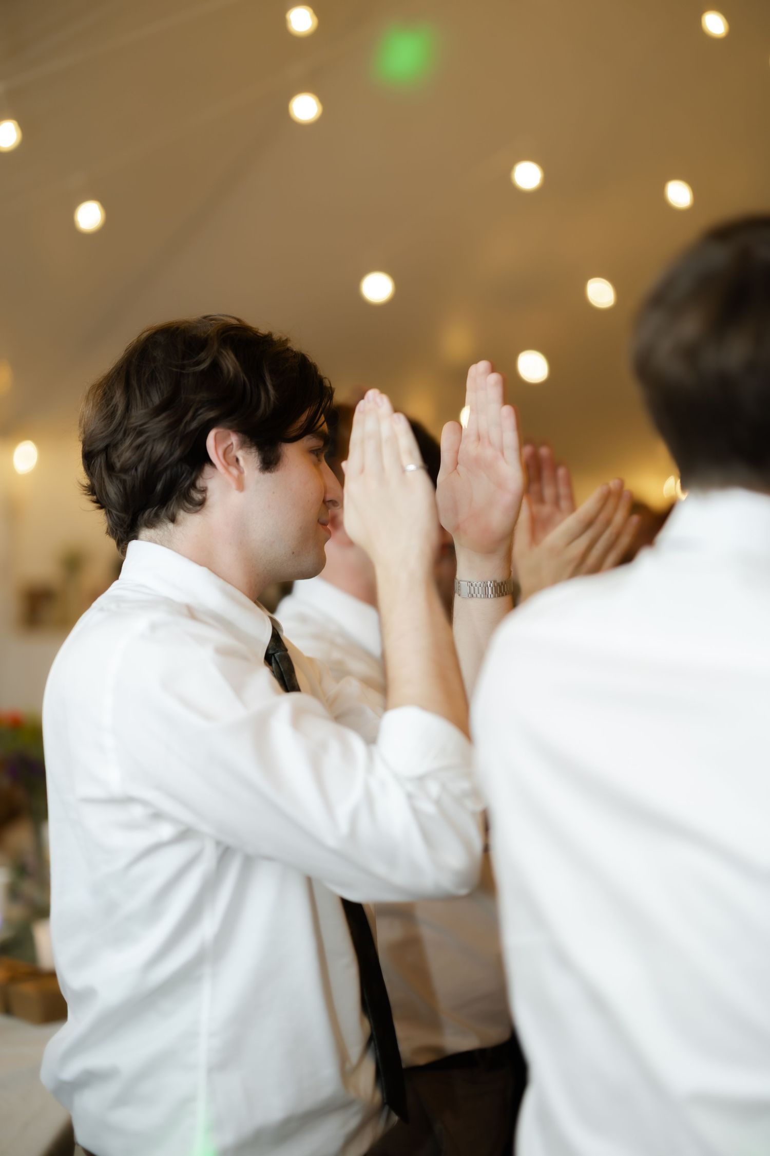 Two men in white dress shirts high-fiving at a festive indoor event with string lights overhead.