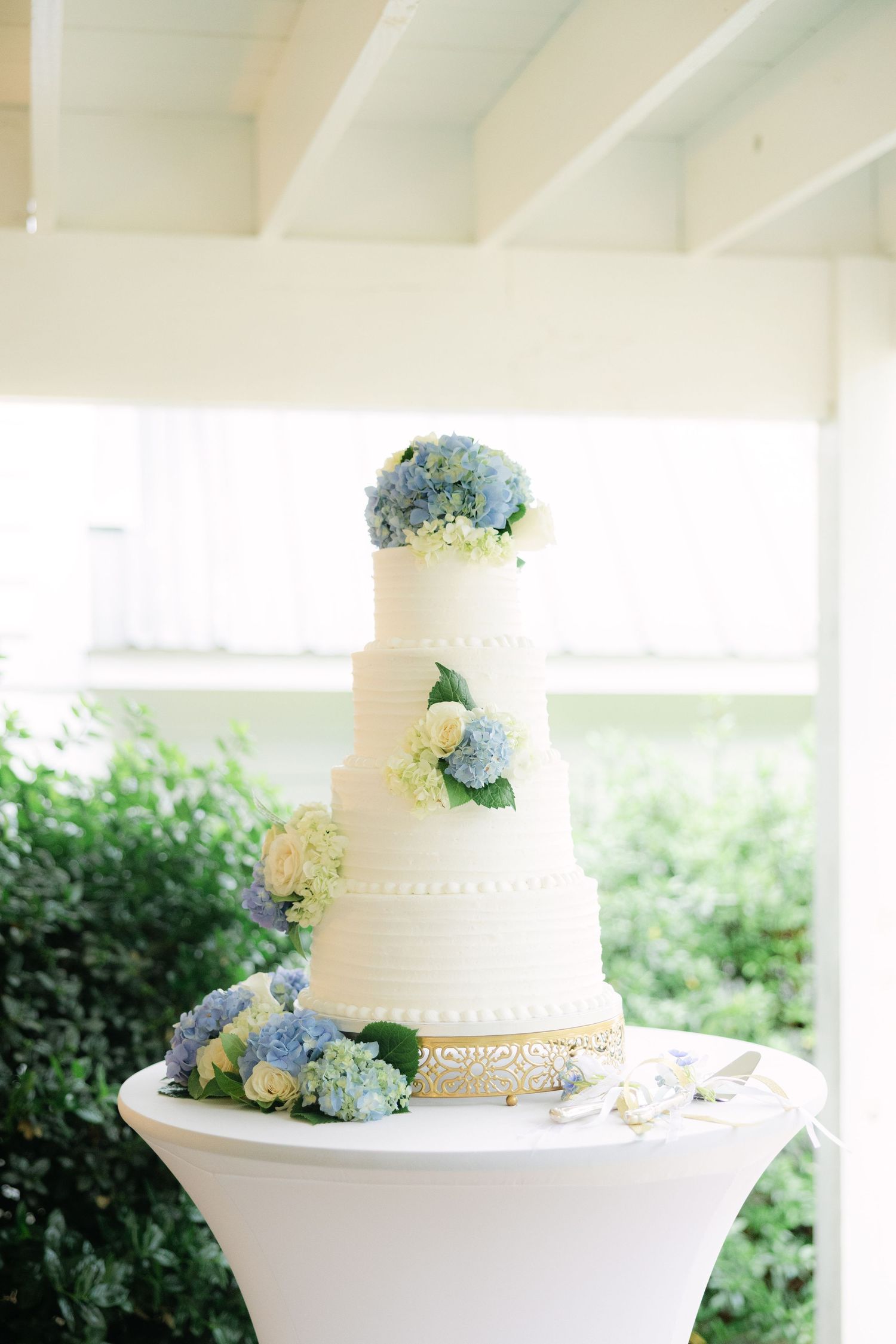 White tiered wedding cake with delicate blue hydrangea and floral decorations displayed on elegant white stand.