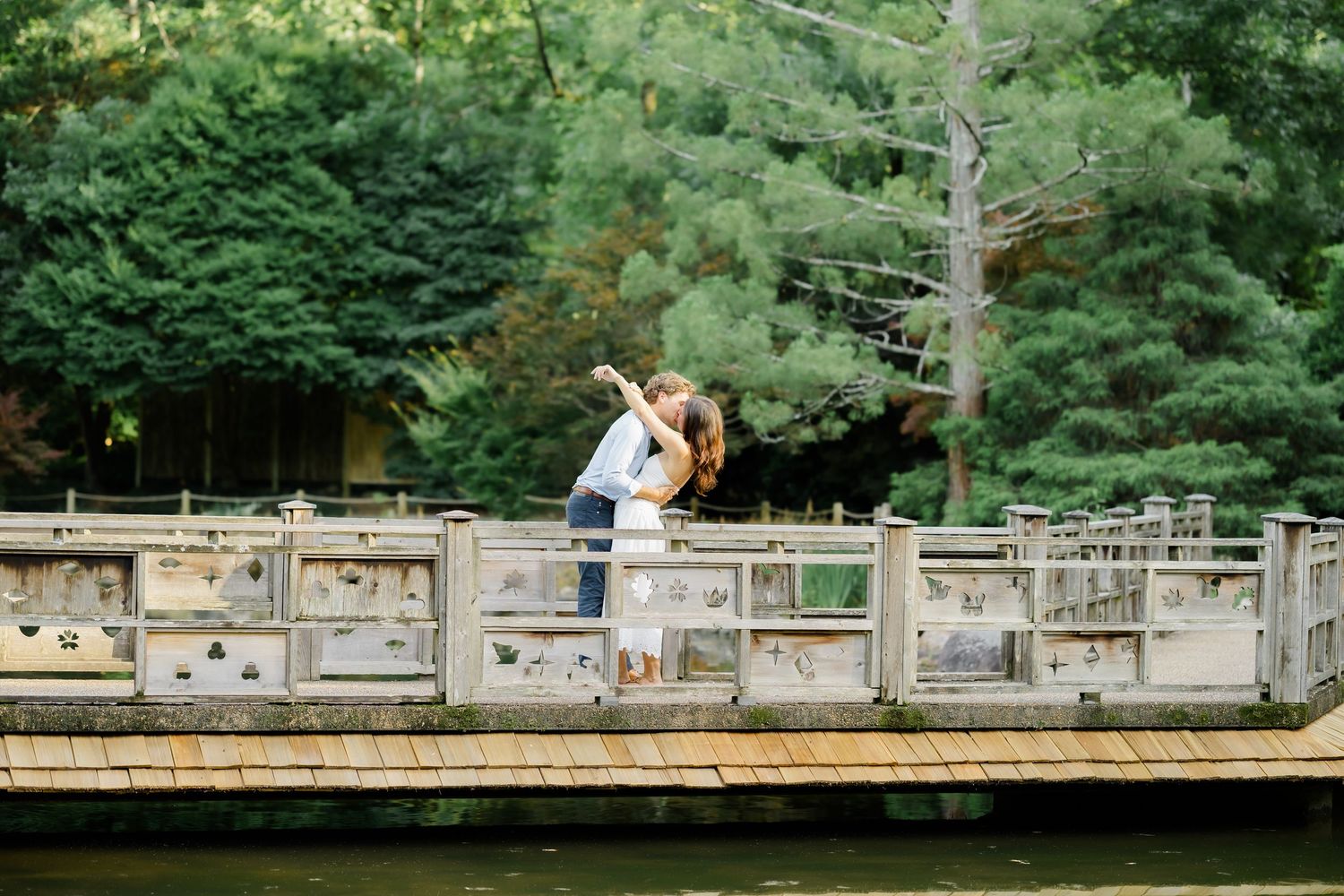 Couple embracing on a stone bridge over a tranquil pond surrounded by lush green trees.