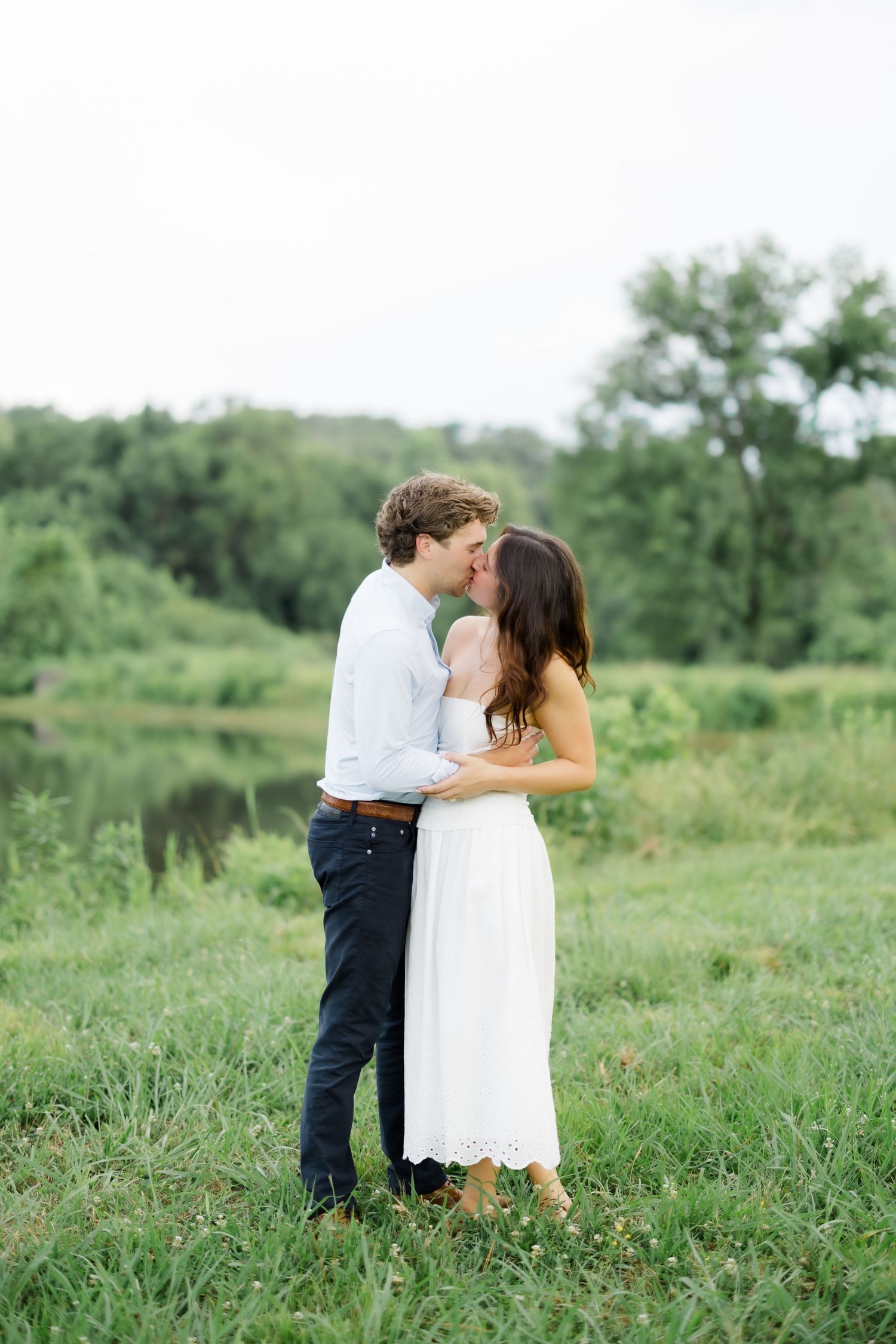 Couple sharing a kiss while embracing in a green meadow near a pond, woman in white eyelet dress and man in blue shirt.