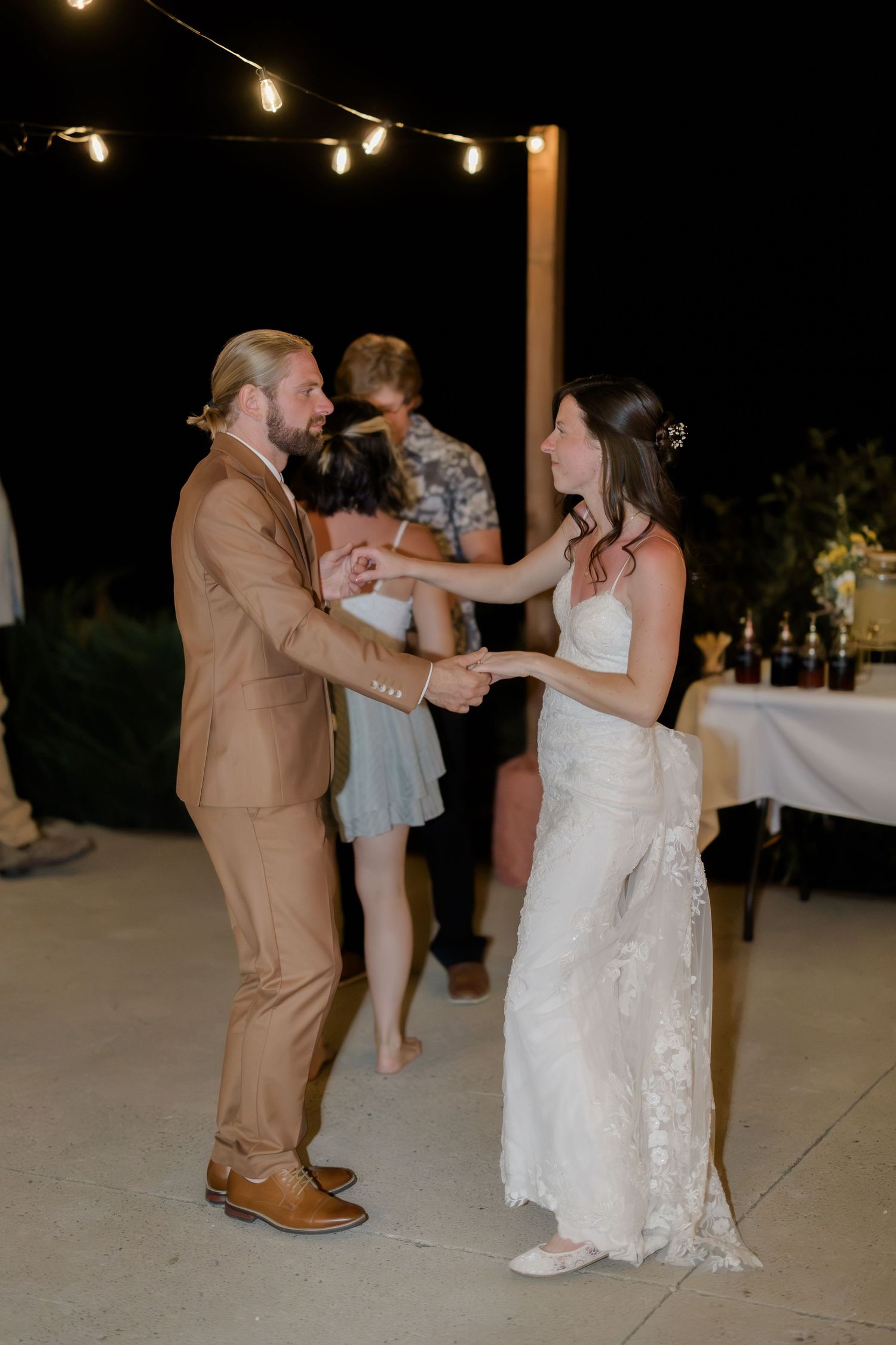 Bride and groom share first dance outdoors at night under string lights, she in lace gown, he in tan suit.