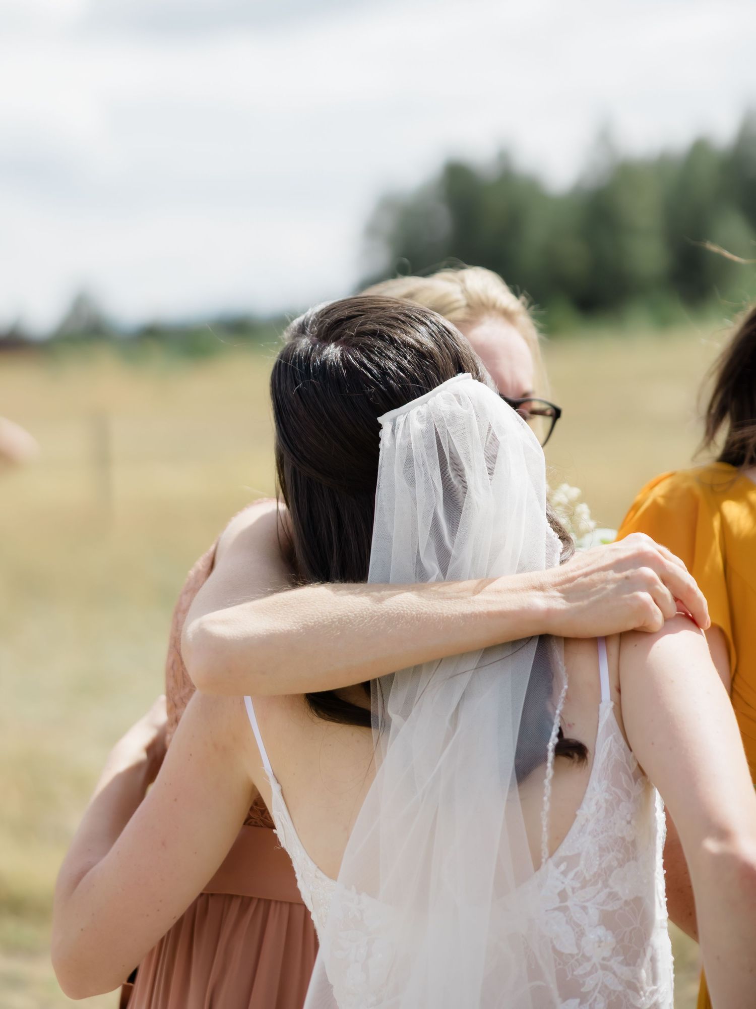 A bride in a white dress and veil shares a heartfelt group hug with guests outdoors at a sunny wedding celebration.