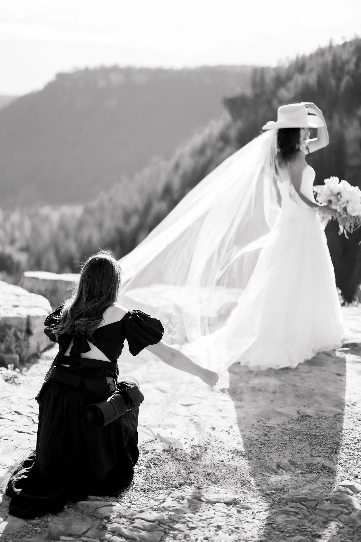 Black and white photo of bride in white gown and cowboy hat with veil flowing, assisted by bridesmaid in black dress outdoors.