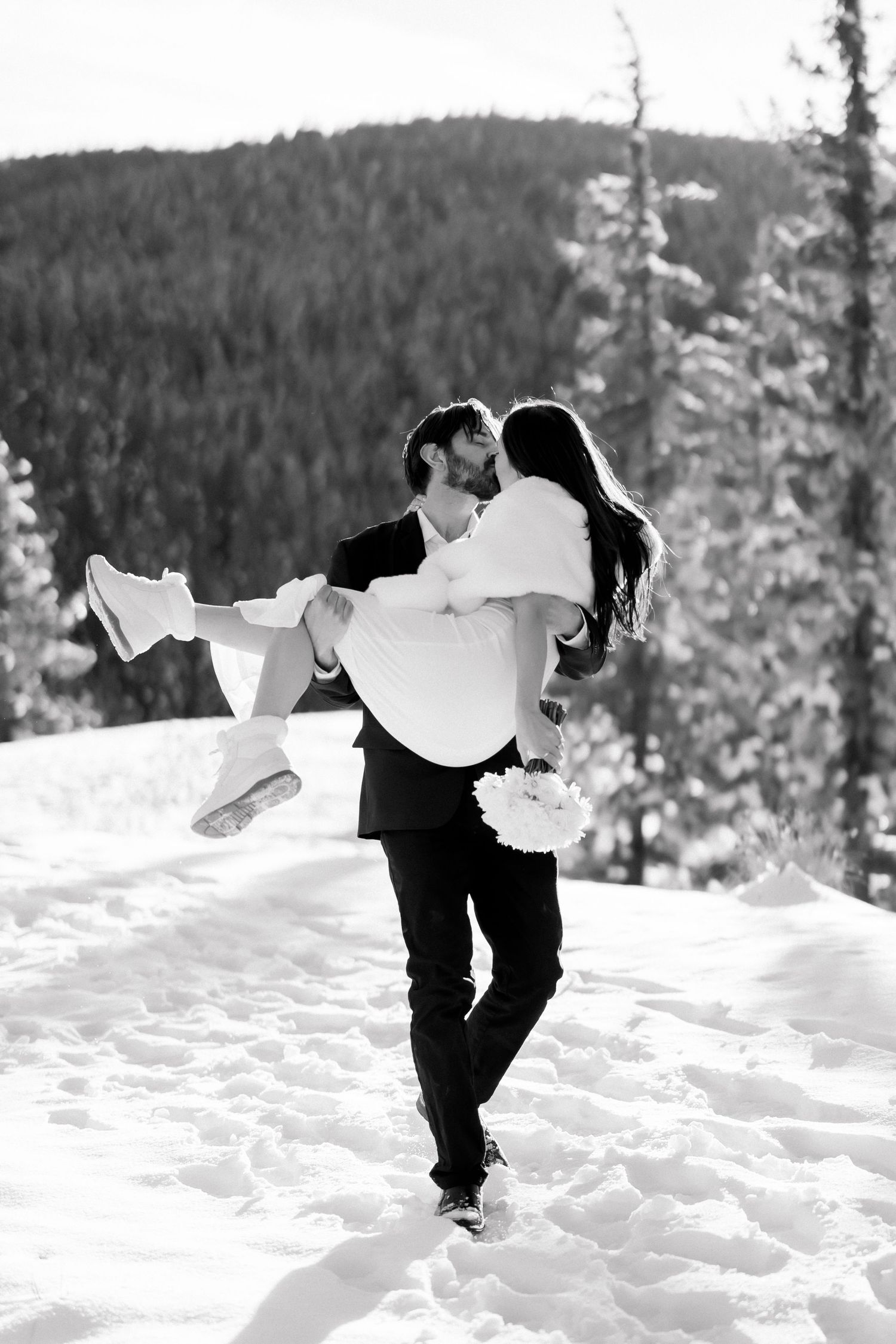 Black and white photo of groom carrying bride in his arms through snowy mountain landscape.