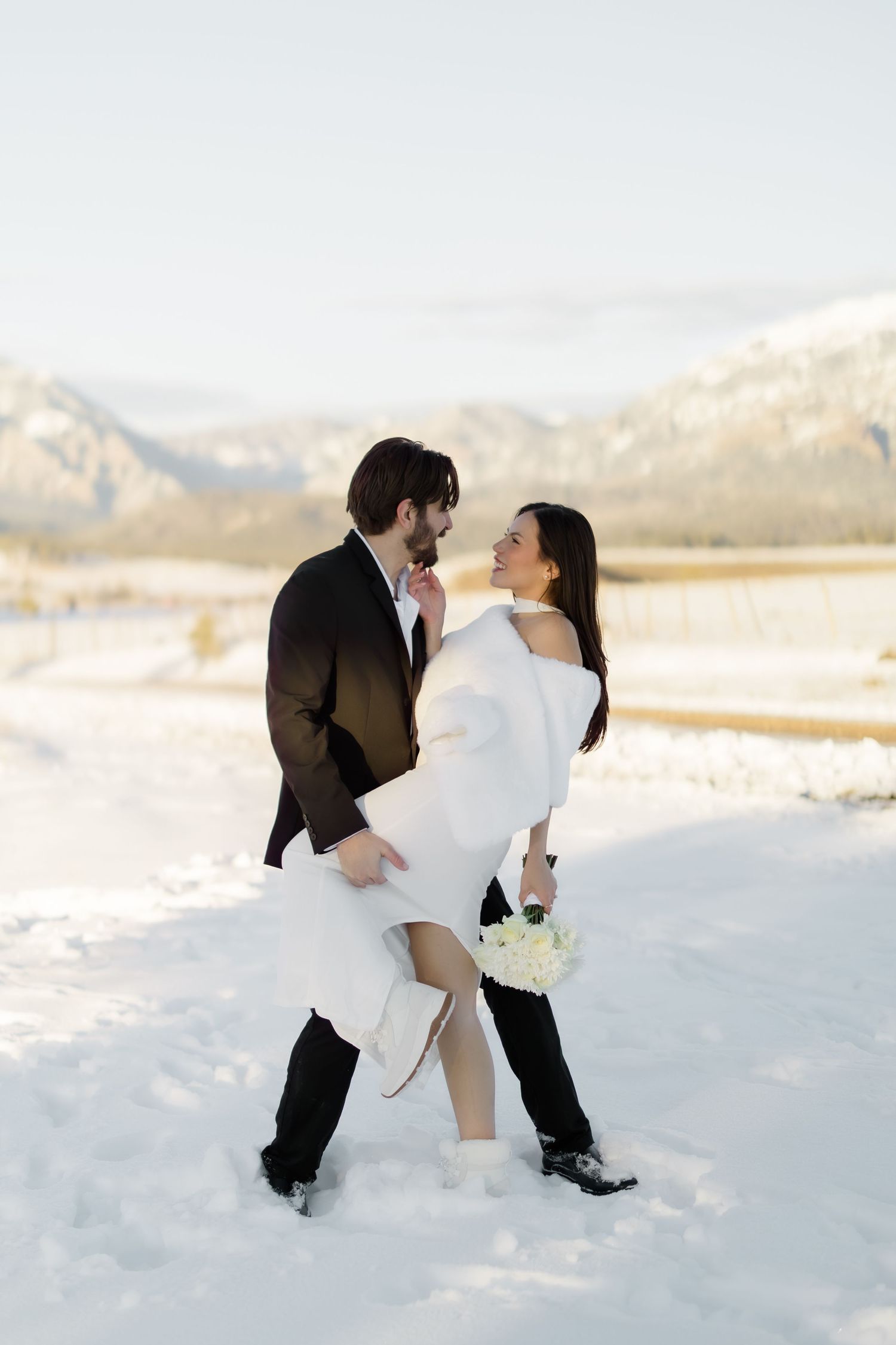 Groom dips bride in white dress and wrap on snow-covered mountain road at sunset.
