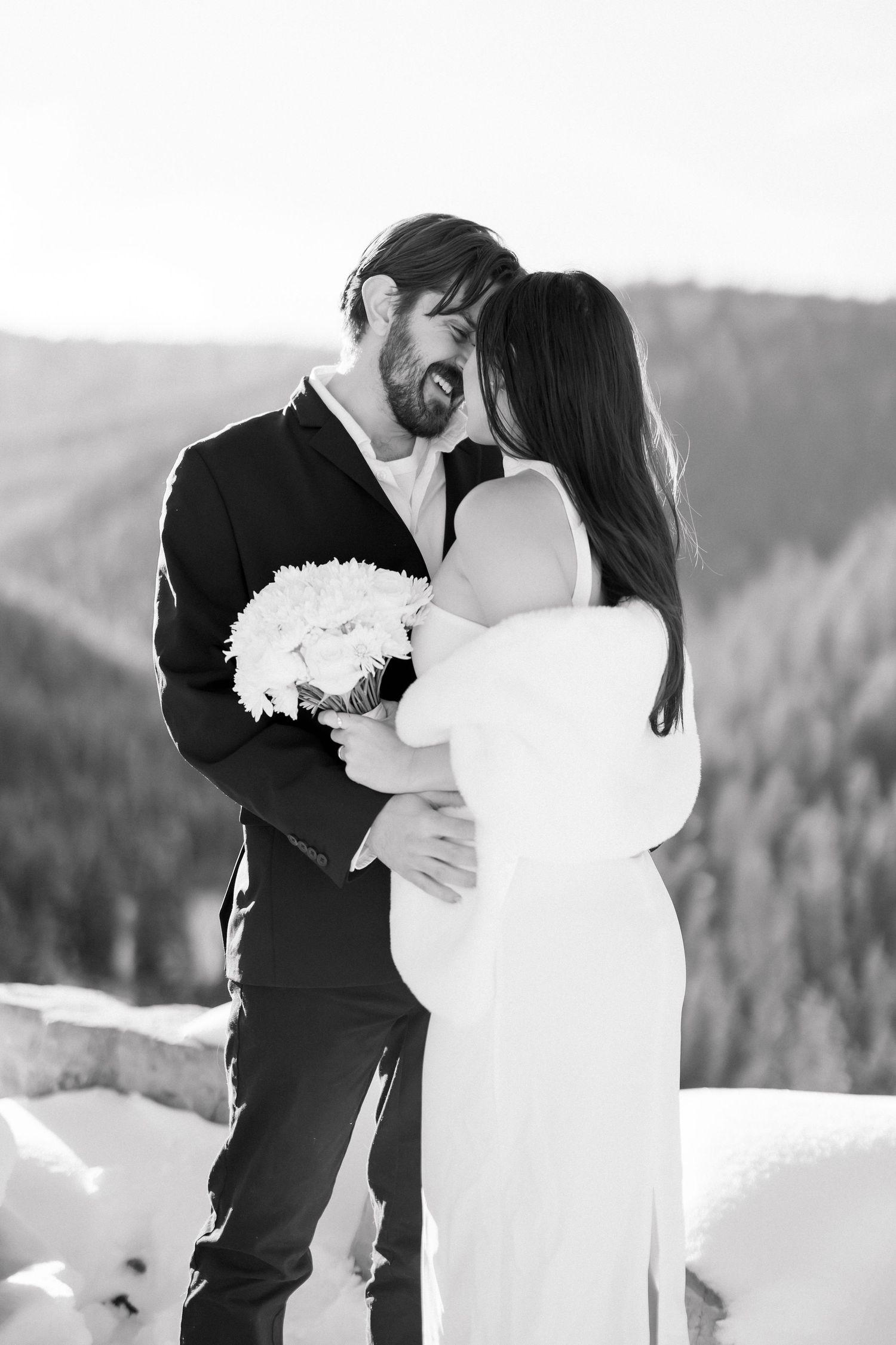 Romantic black and white portrait of bride and groom embracing with bouquet in snowy mountains.