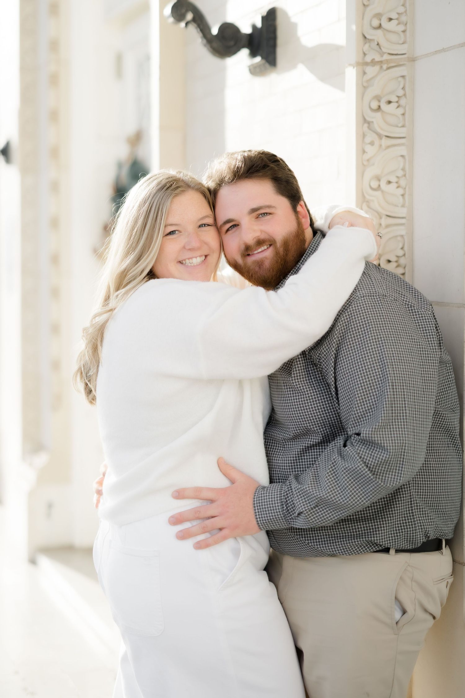 A smiling couple embraces against an ornate white column outdoors during a bright engagement photo session.