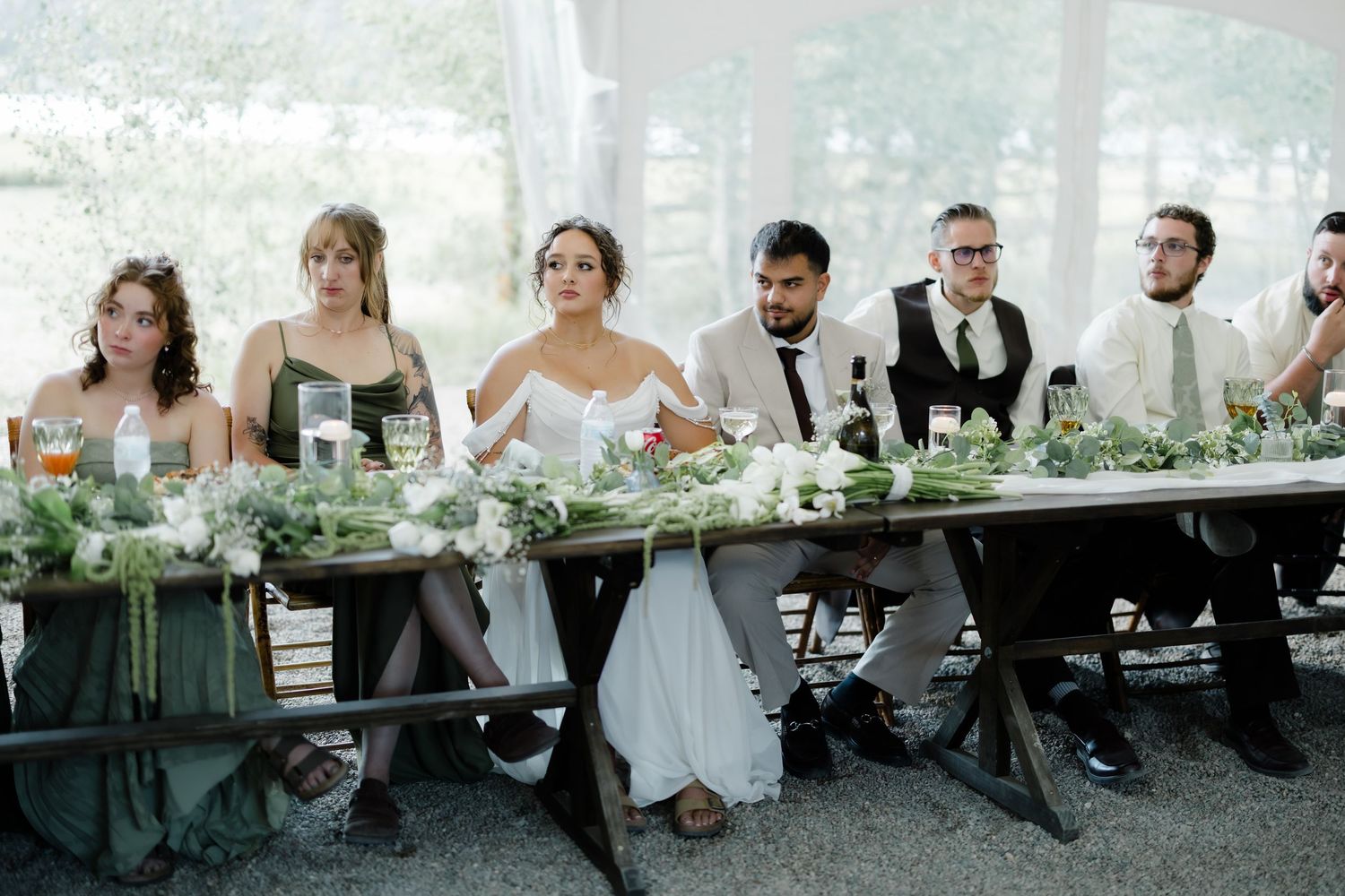 Wedding party seated at long rustic head table with lush greenery and white floral centerpieces.