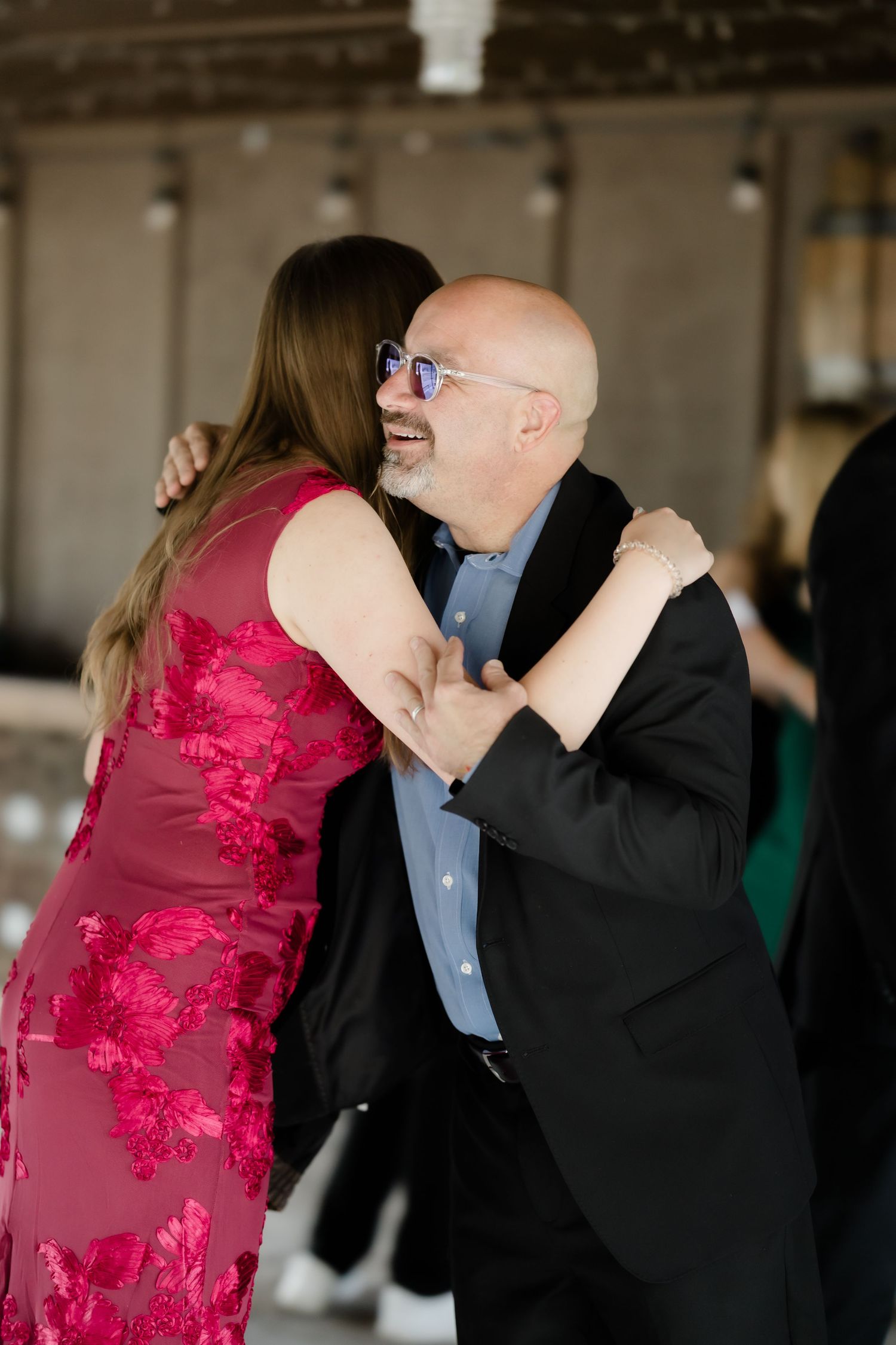 Bald man in black suit with blue sunglasses sharing a warm hug with woman in pink floral dress at an event.