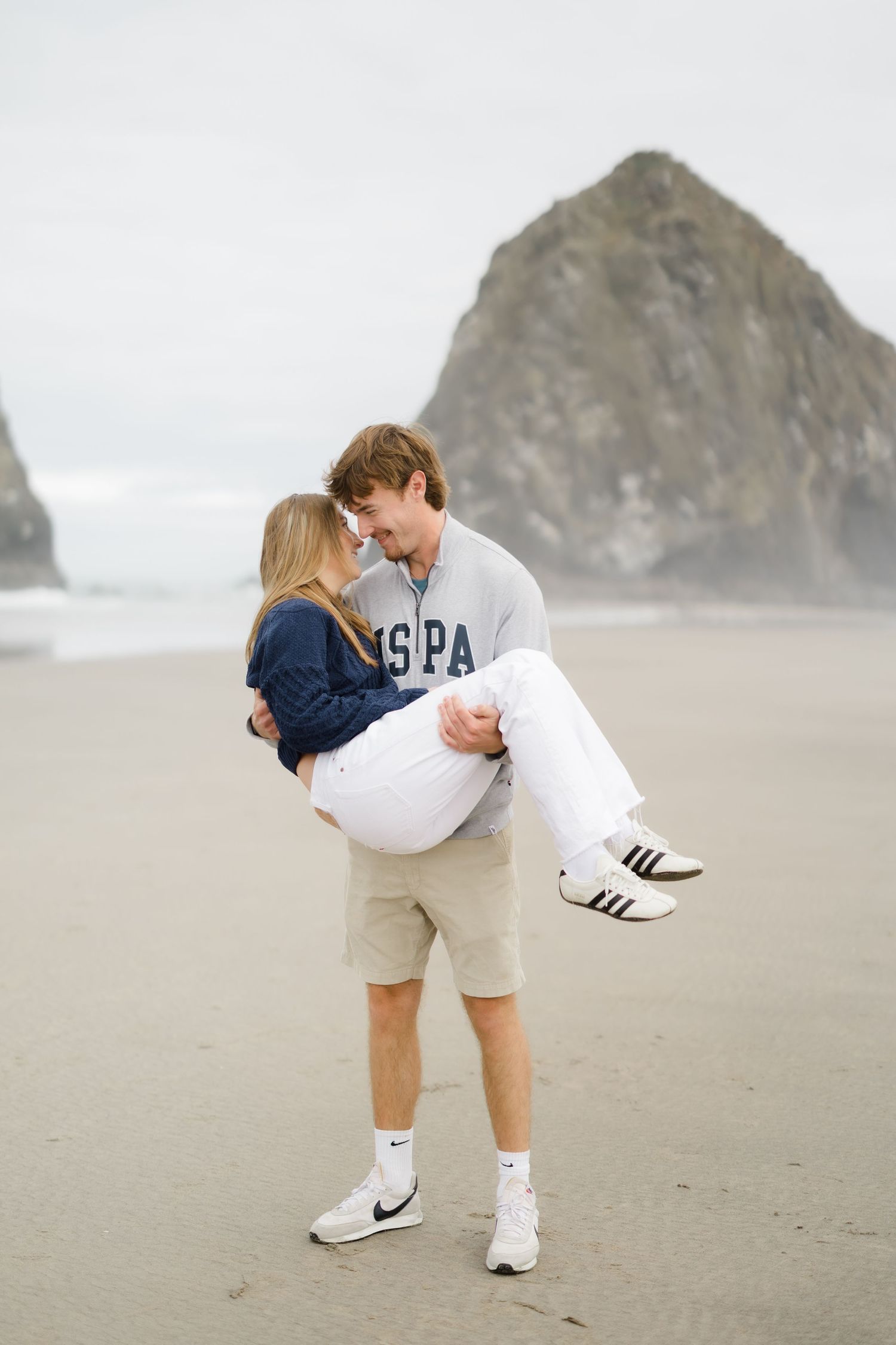 Couple sharing a romantic moment on a sandy beach, man carrying woman in his arms near a large rock formation.