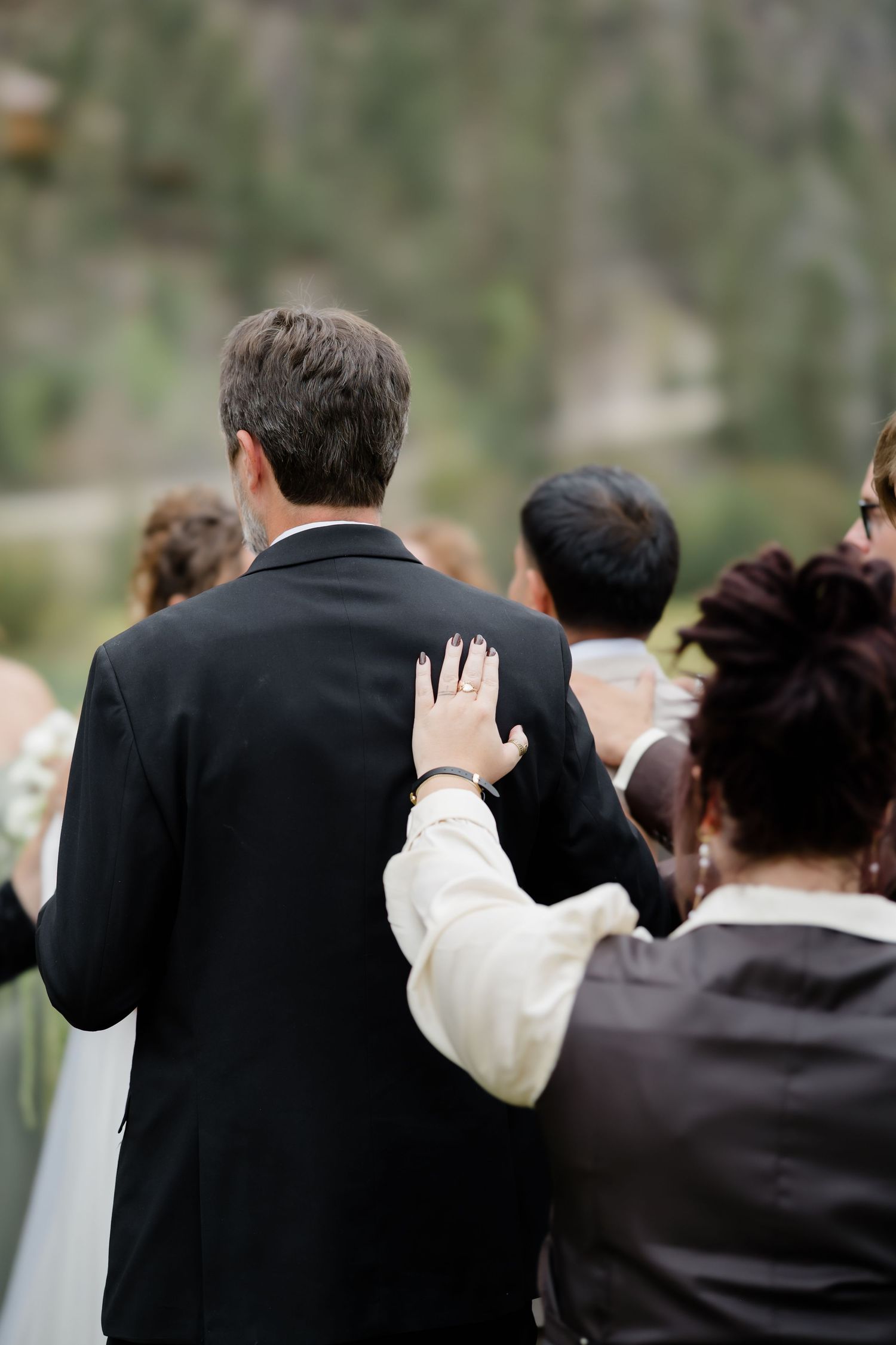 Wedding guests walking together outdoors with a hand resting on a man's shoulder in a black suit.