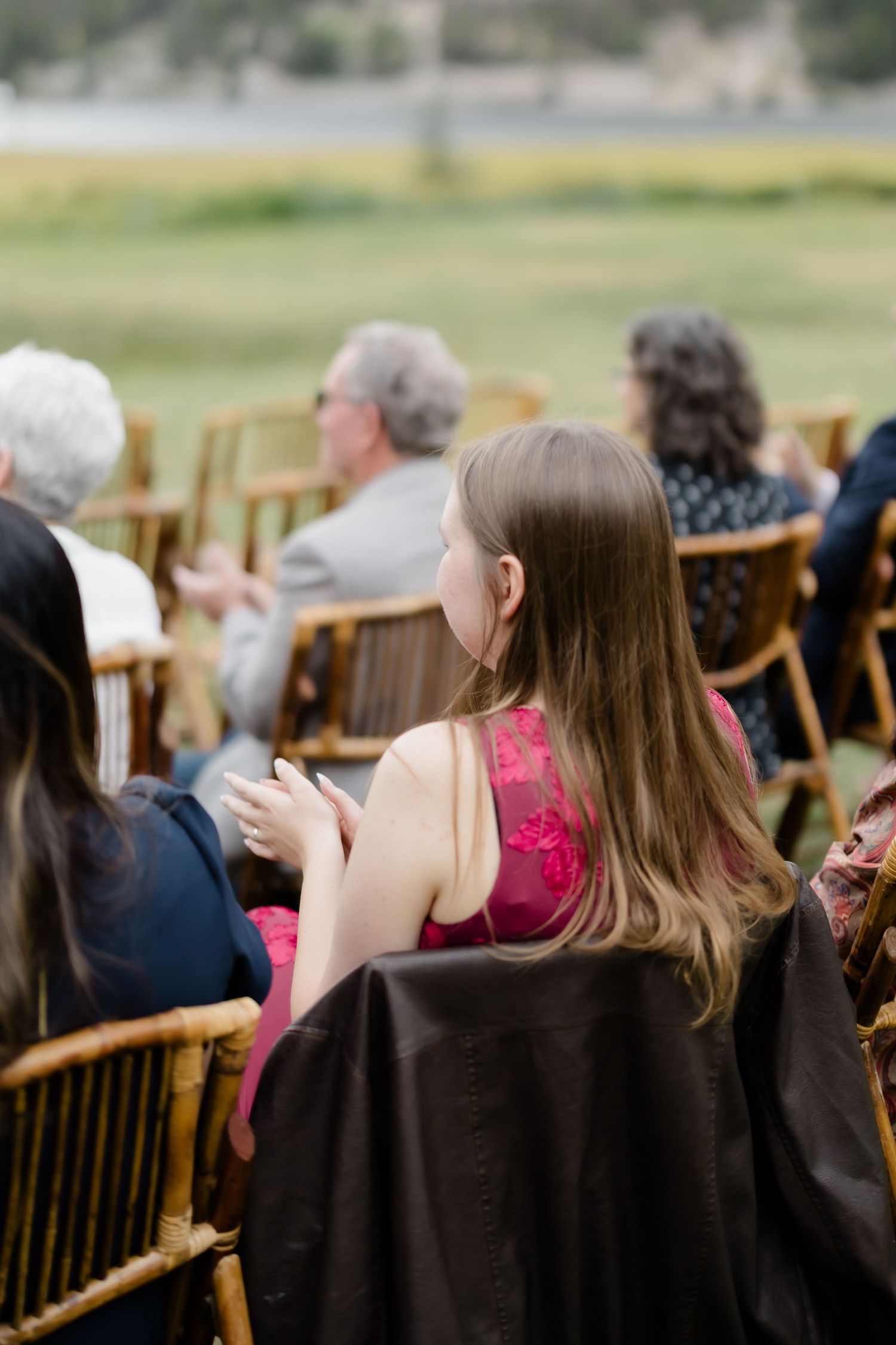 Wedding guest in pink dress seated in bamboo chair at outdoor ceremony with green meadow backdrop.