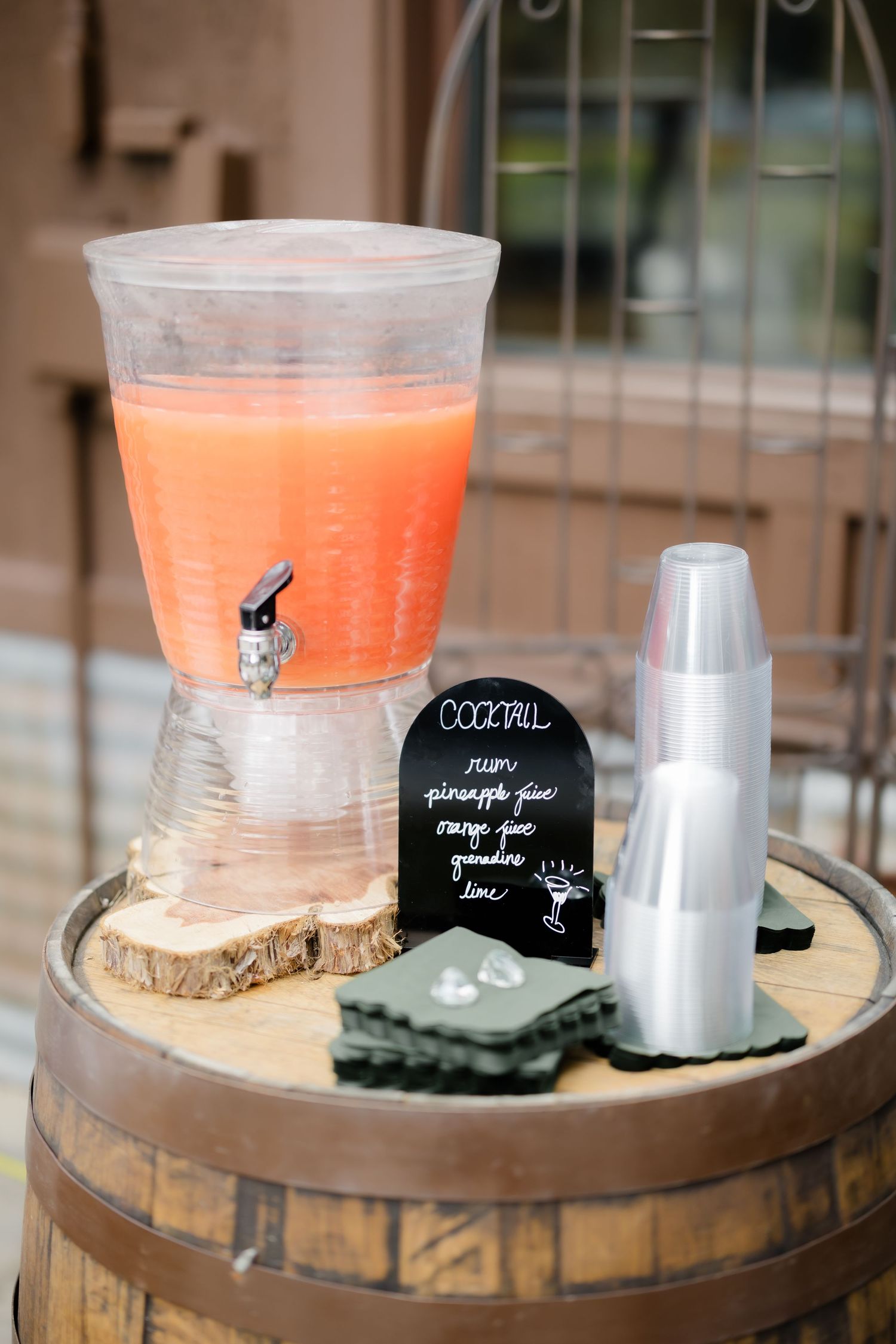 A large beverage dispenser filled with orange cocktail punch sits on a rustic wooden barrel with cups and a chalkboard sign.