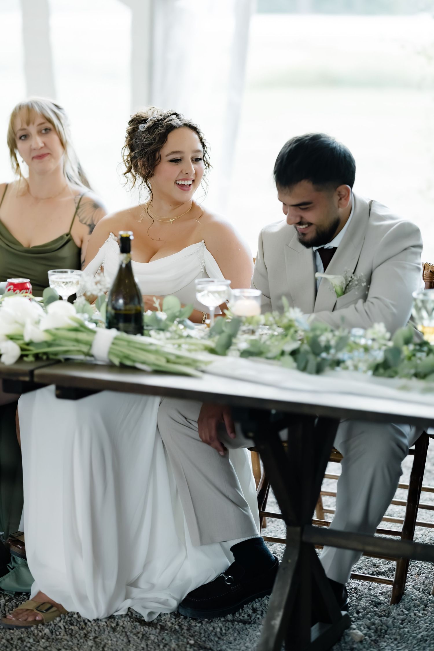 Bride and groom laughing together at sweetheart table with floral greenery runner during wedding reception.