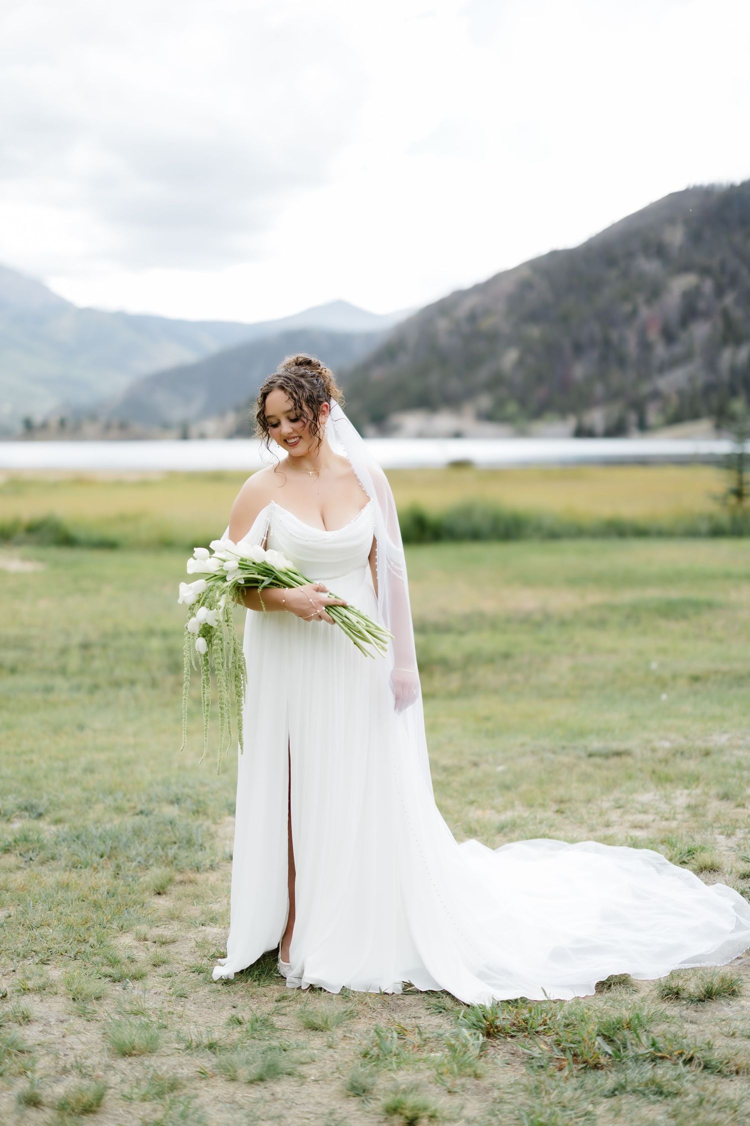 Smiling bride in white gown with veil holds white floral bouquet in scenic mountain setting.